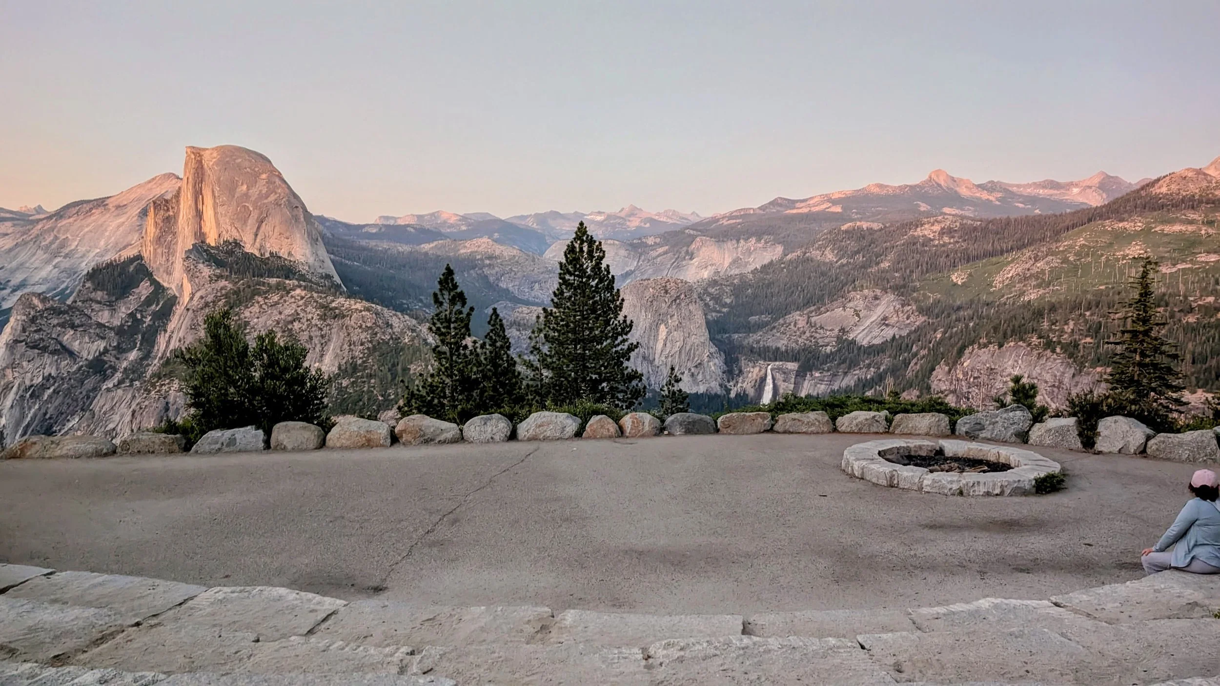 Mountain landscape with large granite dome, forested slopes, and distant mountain peaks during sunset. In the foreground, there is a paved area with rocks and a person sitting near a fire pit.