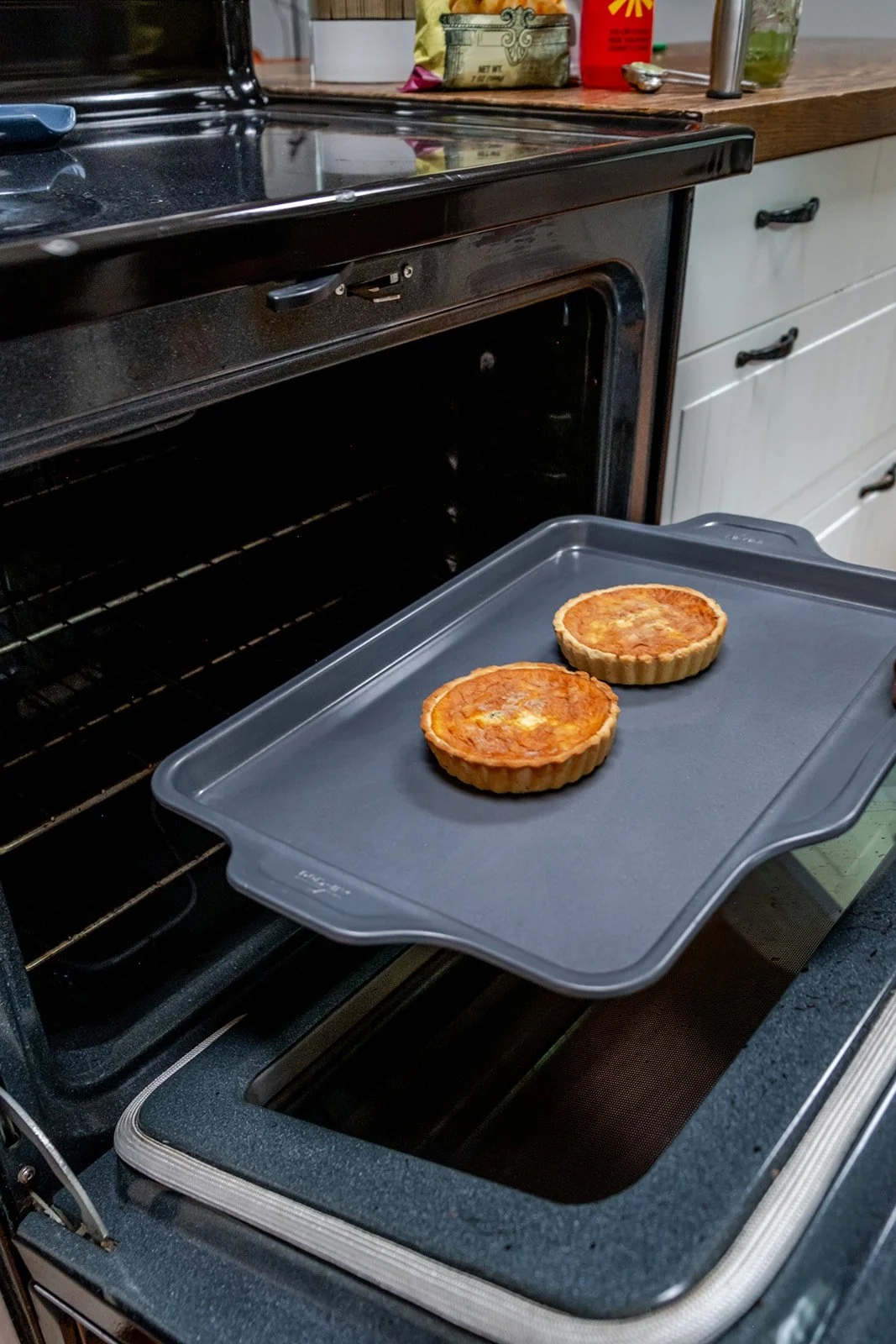 Two egg tarts cooling on a gray baking sheet in an oven, with kitchen counters and cabinets in the background.