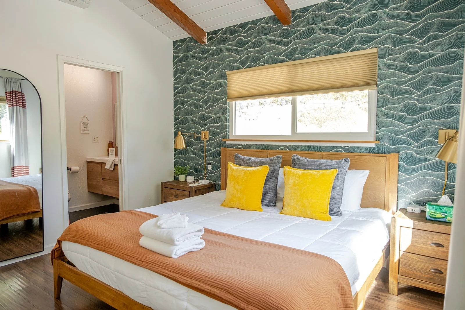 A bedroom with a wooden bed frame, white bedding, and four pillows (two yellow and two gray). There are two wooden nightstands with lamps, a potted plant, and a small white object on one nightstand. Behind the bed, there's a blue wave-patterned accen