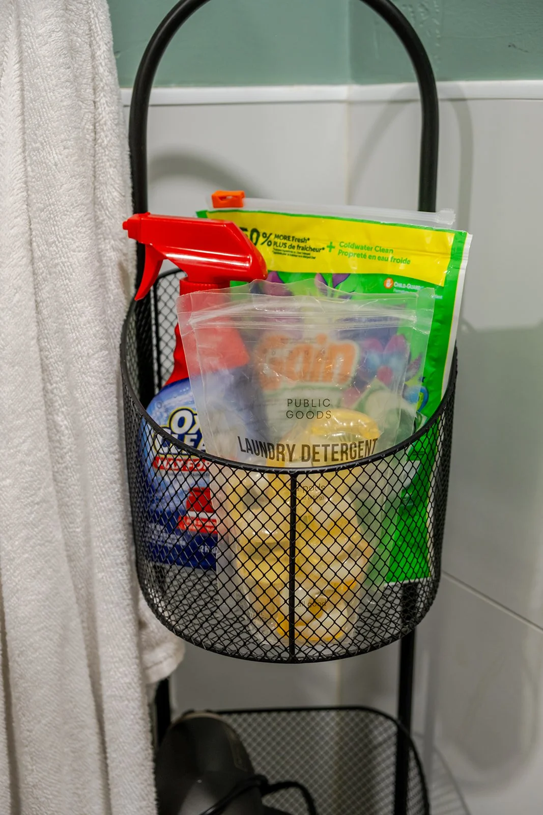 Black metal hanging organizer with cleaning supplies and snacks, placed against a bathroom wall near a towel and white tile