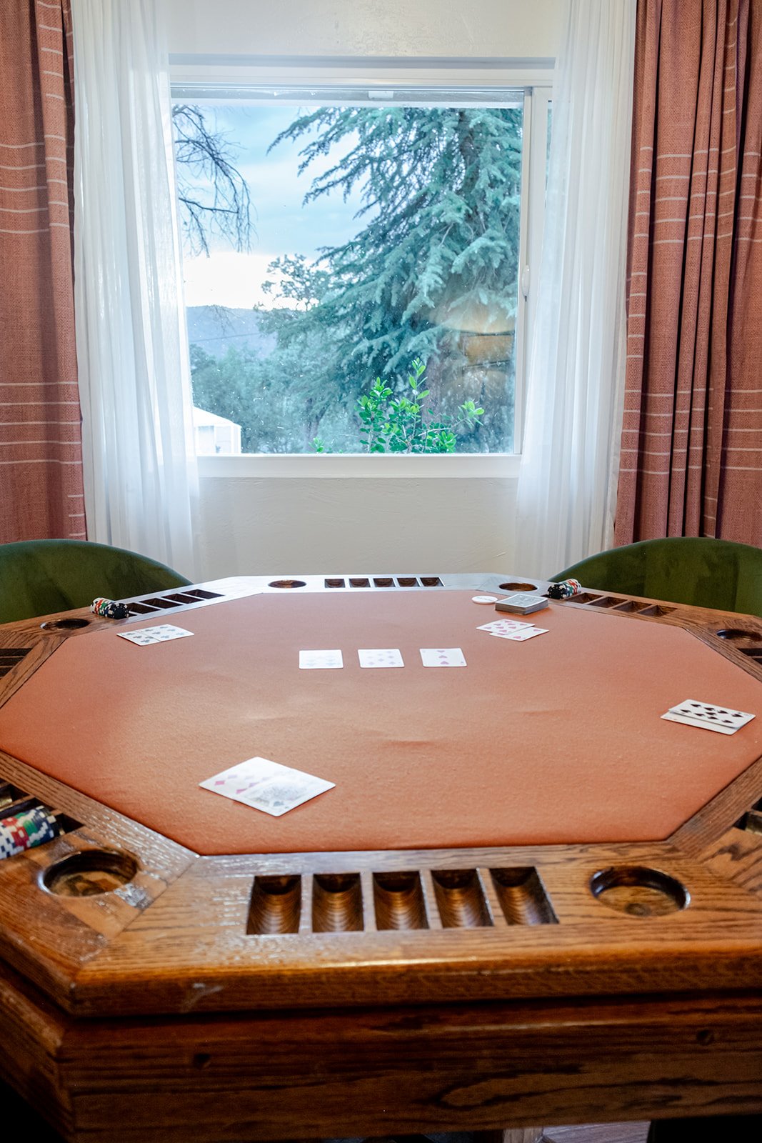 A poker table with cards and chips inside a room with a window showing snow-covered trees outside.