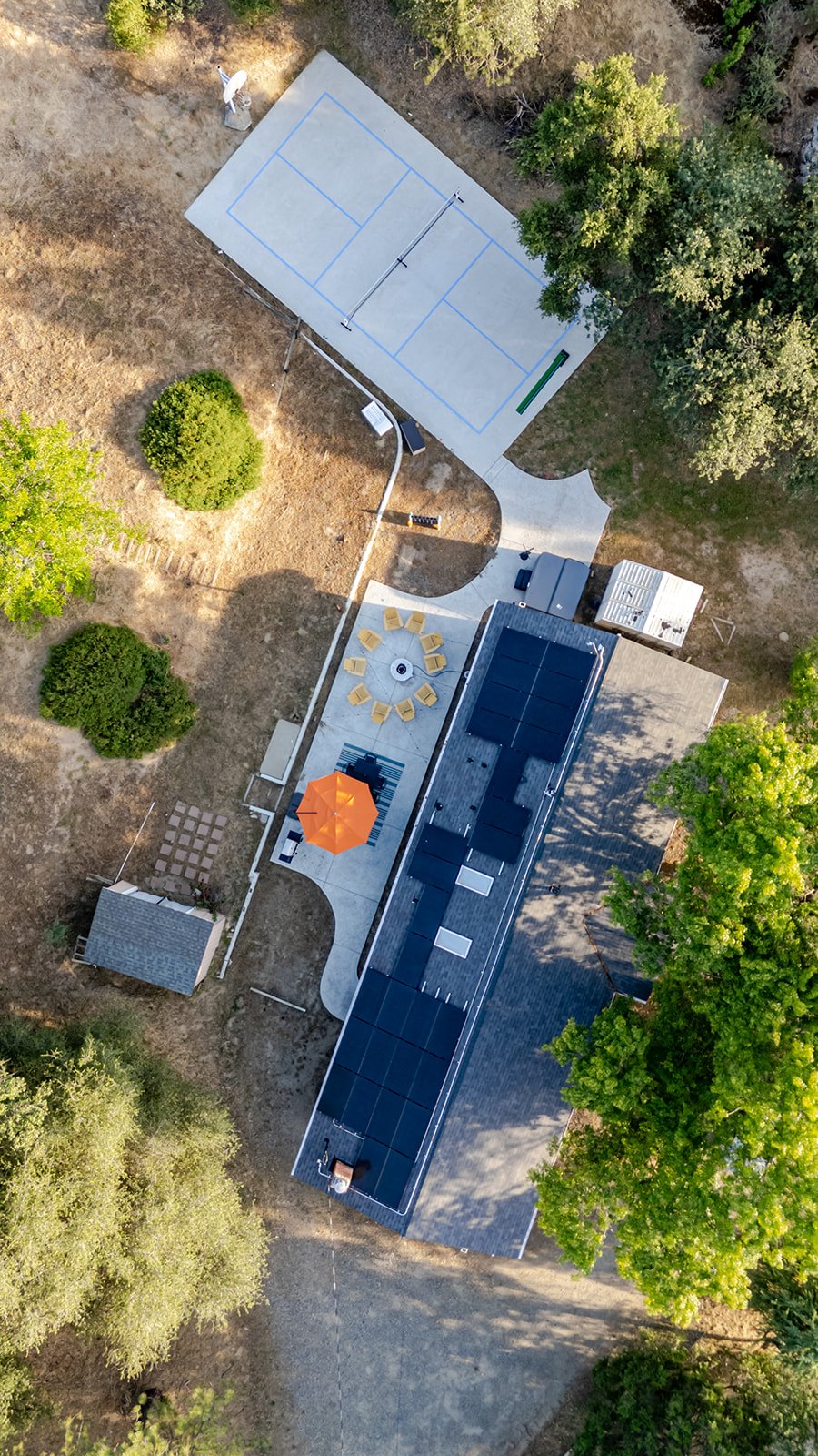 Aerial view of a backyard with a tennis or basketball court, patio with yellow chairs around a fire pit, an orange umbrella, and solar panels on the roof of a house, surrounded by trees and shrubs.