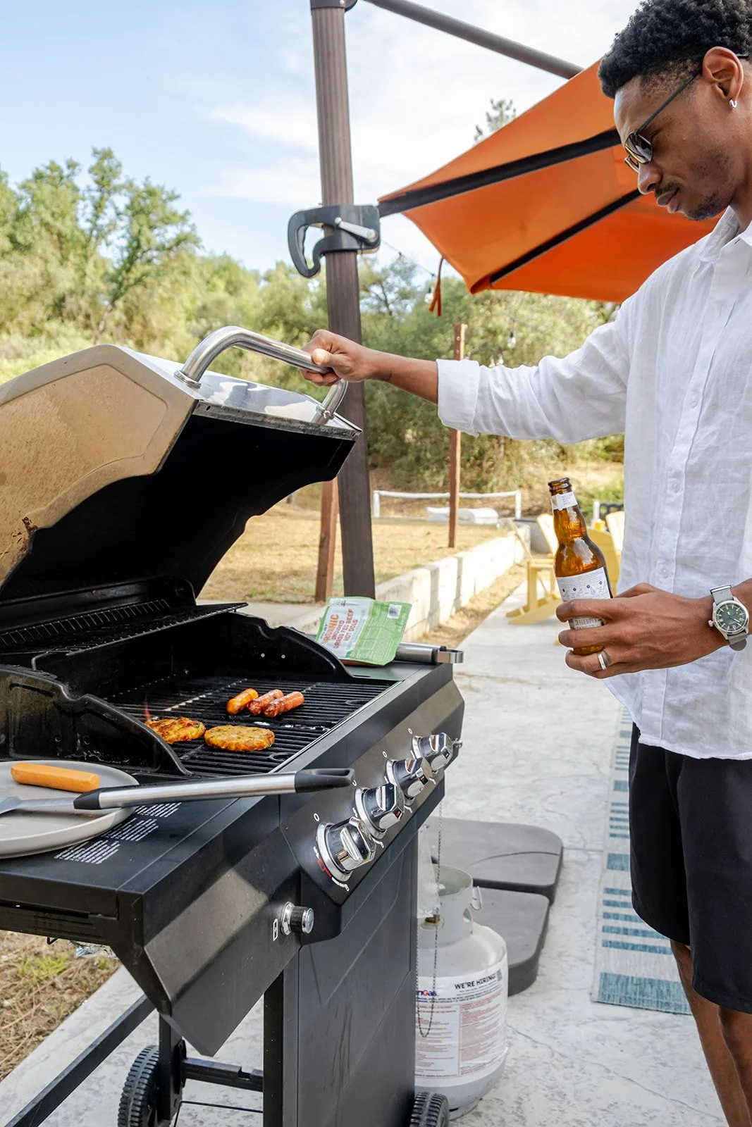 A man grilling hot dogs and burgers on an outdoor barbecue grill, holding a beer bottle, with a garden and outdoor seating area in the background.
