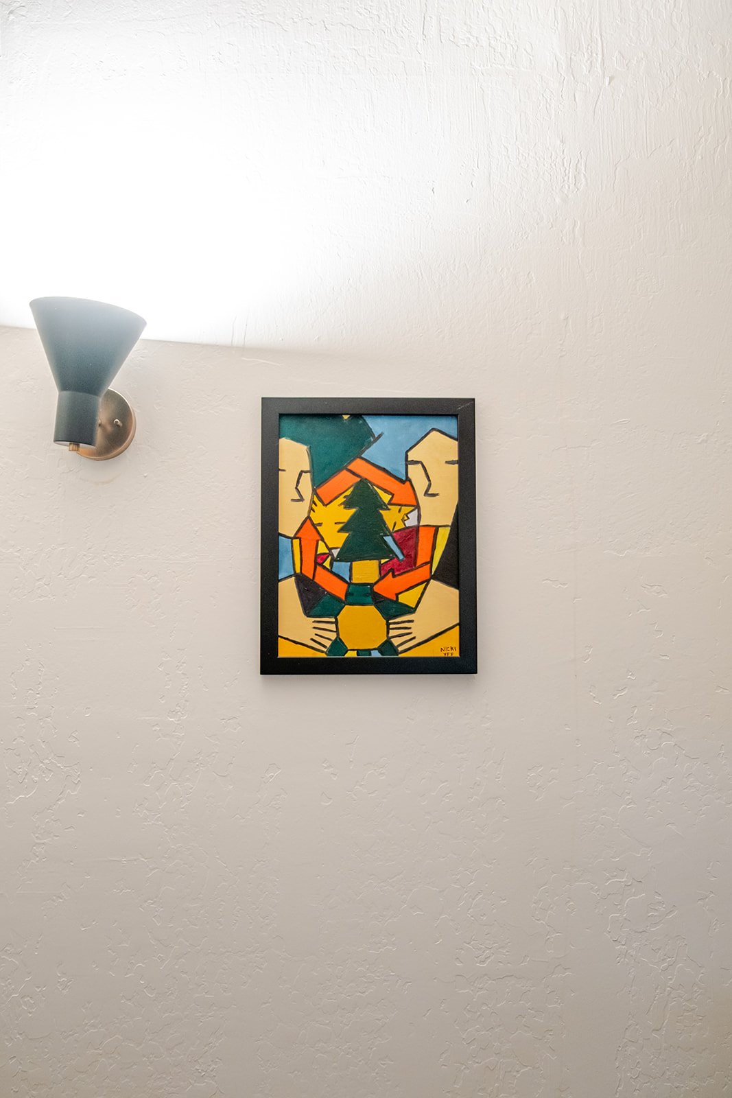 Colorful abstract art featuring a black-framed painting of two people holding hands and an evergreen tree in the center, mounted on a white wall with a wall-mounted light fixture nearby.
