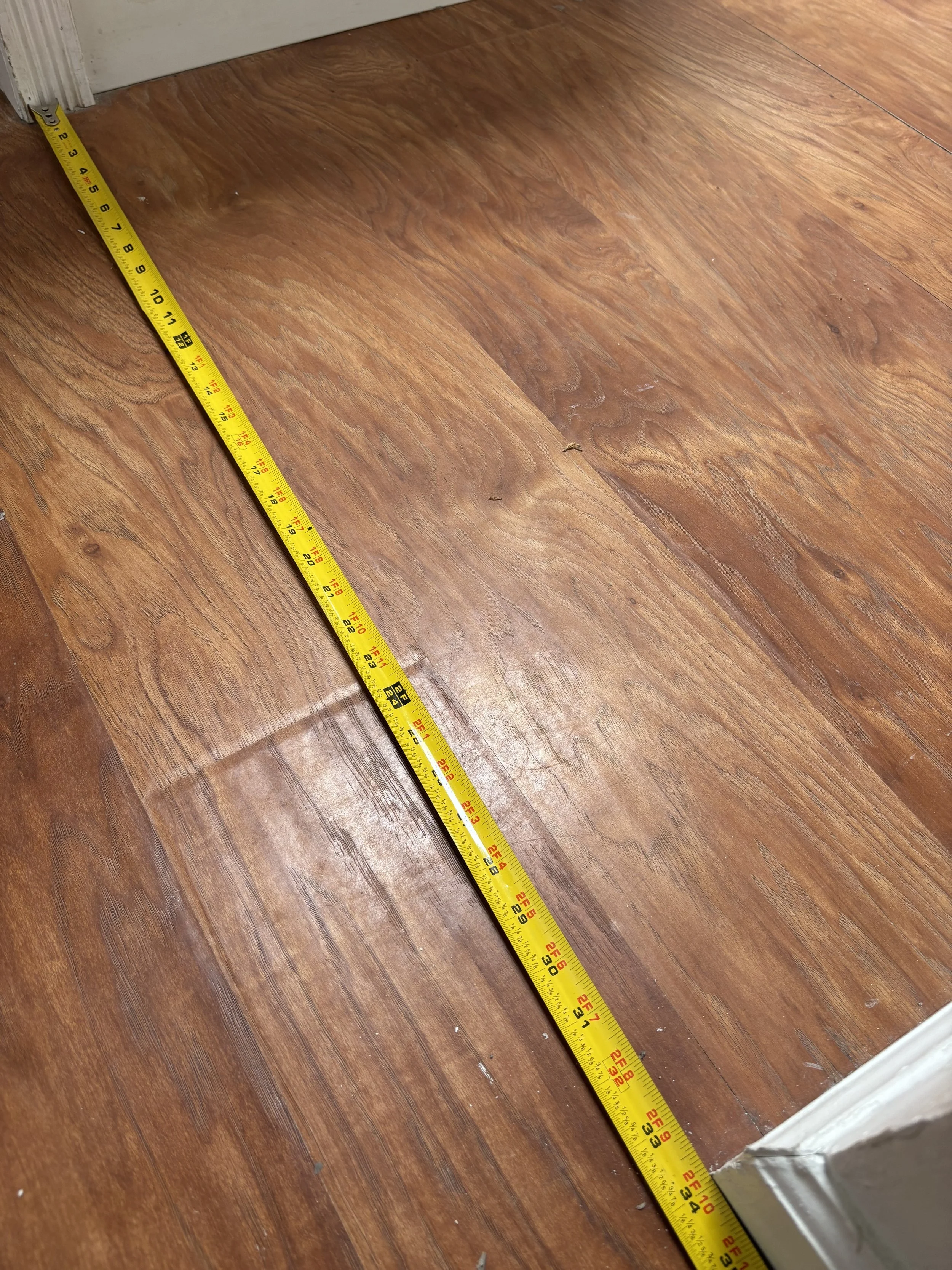 A wooden floor with a yellow measuring tape stretched across it, starting at the base of a door frame near the bottom left corner of the image.
