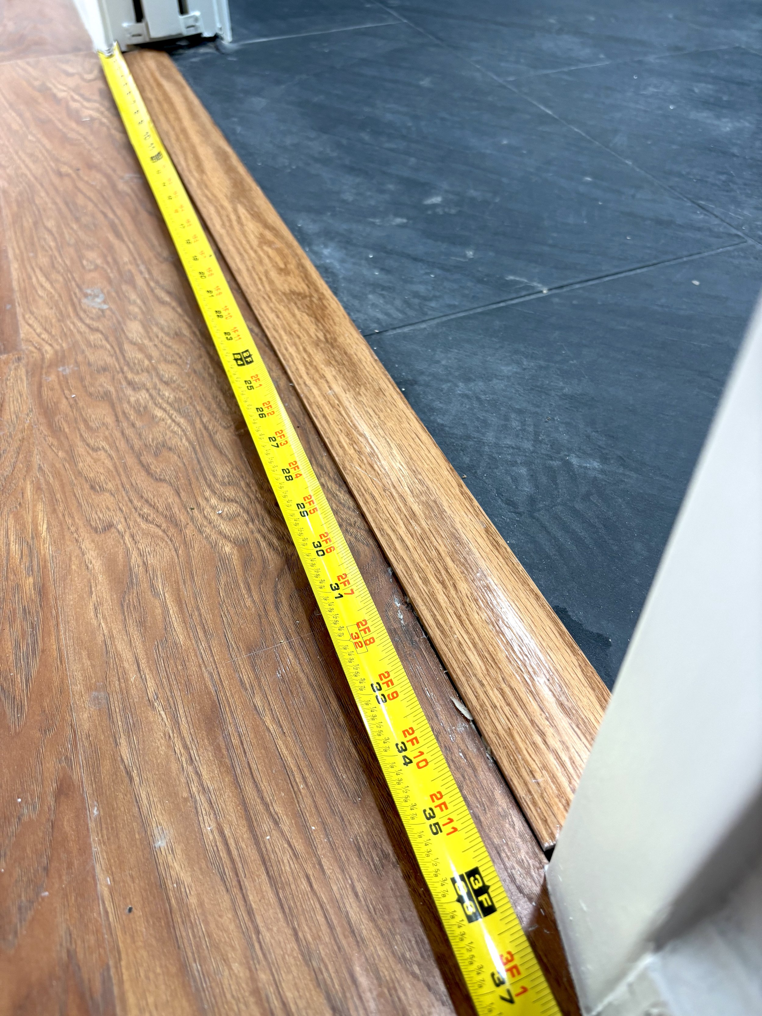 A yellow measuring tape laid along the edge of a wooden floor, extending across a transition to a dark tiled floor in a room.