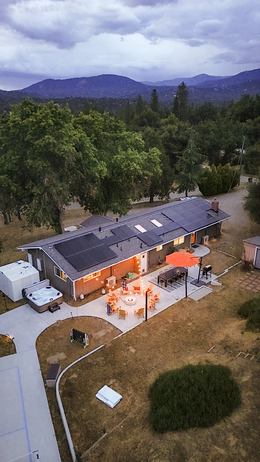 A house with solar panels on the roof, a backyard patio with a fire pit, chairs, tables, an umbrella, and string lights, surrounded by trees and mountains in the background at dusk.