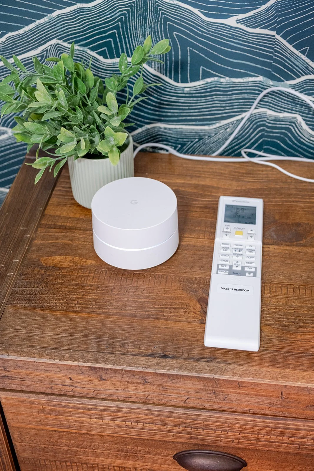 A wooden nightstand holding a potted green plant, a white Google Nest device, and a white remote control with labeled buttons. The background features a patterned blue wall with white and darker blue lines.