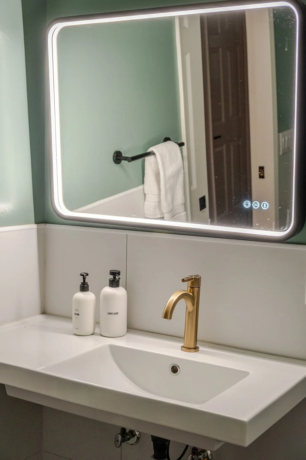 Bathroom sink with a gold faucet, two white soap dispensers labeled lotion and hand soap, a large mirror with LED lighting, and a towel hanging on a black towel bar.