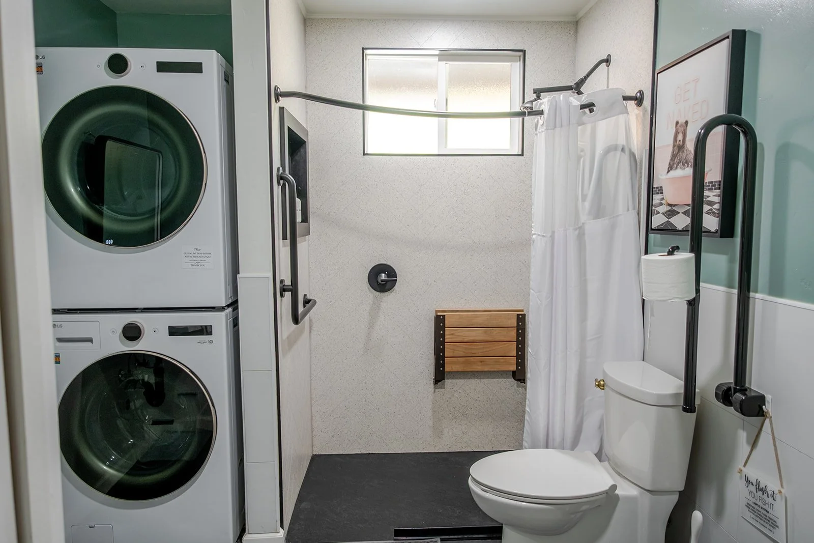 A bathroom with a laundry area with a stacked washer and dryer on the left, a window at the back, a shower curtain, a toilet with grab bars, a small wooden bench, and a framed picture on the wall.