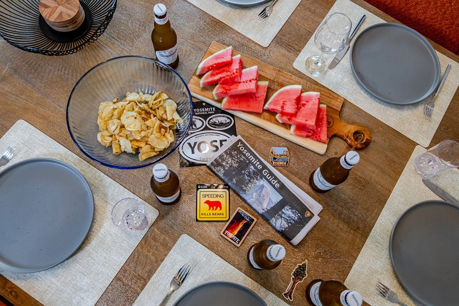 A table set for a meal with gray plates, forks, knives, and water glasses. In the center, there is a watermelon on a wooden cutting board, a large bowl of potato chips, and several small bottles of beer. There are items related to Yosemite National P