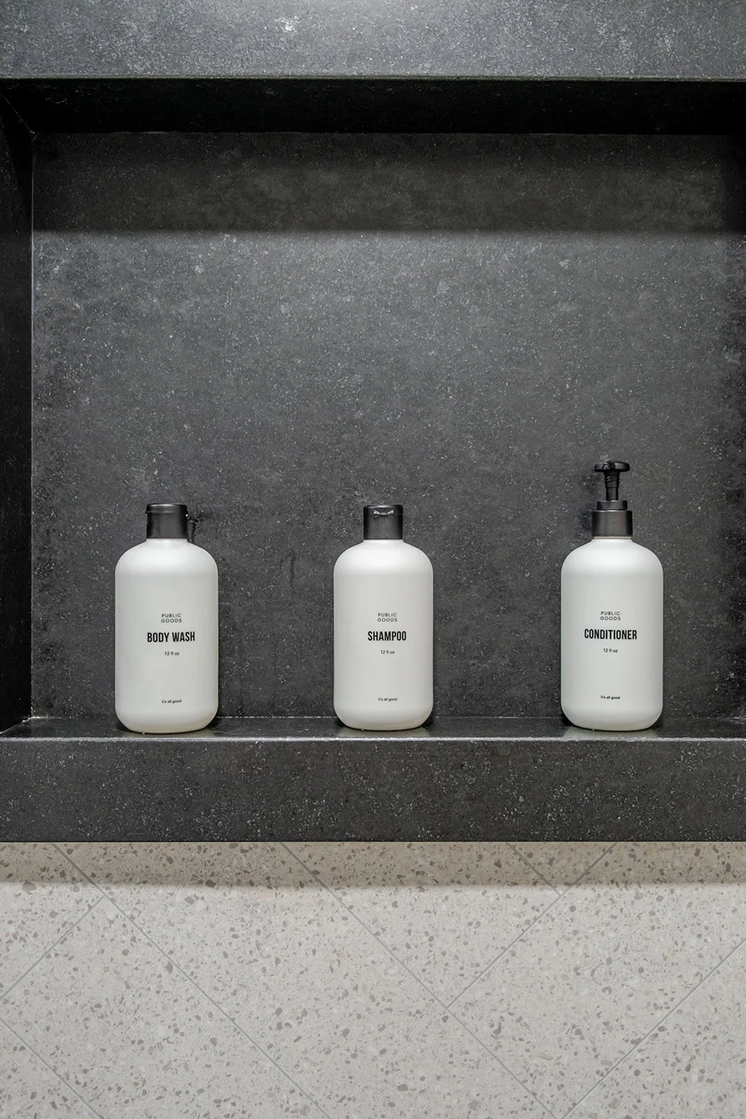 Three minimalist white bottles labeled 'Body Wash', 'Shampoo', and 'Conditioner' on a black shelf against a dark gray wall.
