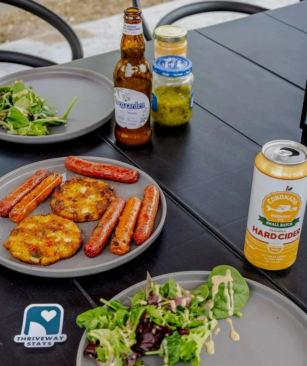 Outdoor table with plates of salad, grilled vegetables, and sausage, along with jars of pickles and mustard, a bottle of beer, and a can of pineapple hard cider.