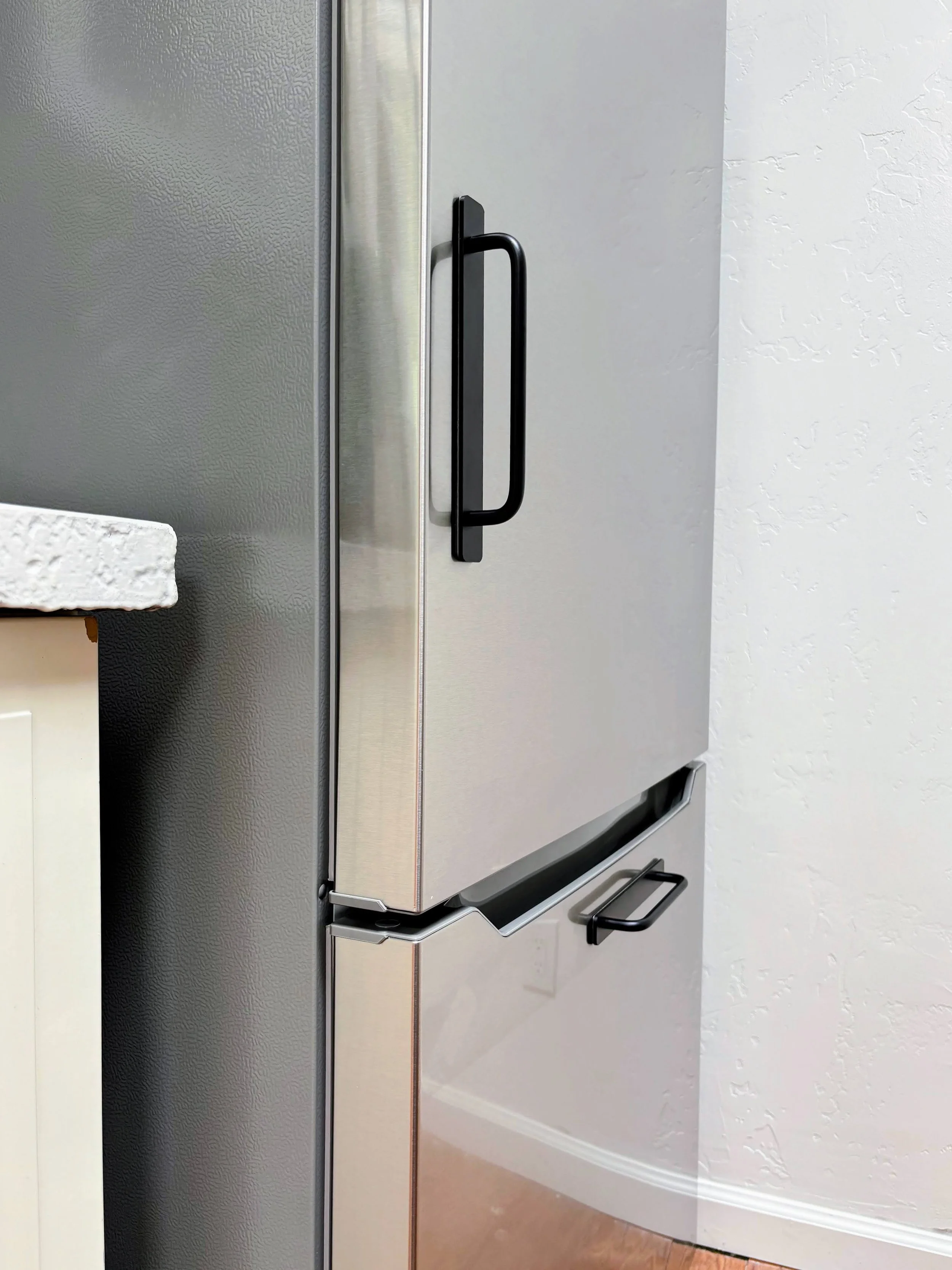 Close-up of a stainless steel refrigerator door with a black handle in a kitchen. Part of a white countertop and textured wall is visible.