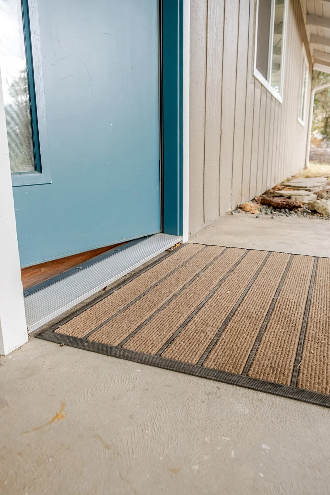 Close-up of a light blue front door with a metal threshold, on a concrete porch with a brown doormat. Part of the house's beige wooden siding and a window beside the door are visible, with a pathway leading away from the house.