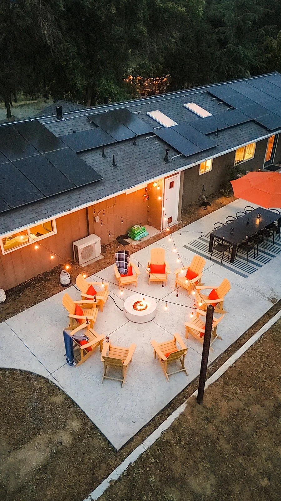 A backyard patio with outdoor seating arranged in a circle around a fire pit, string lights hanging overhead, and a house with solar panels on the roof in the background.