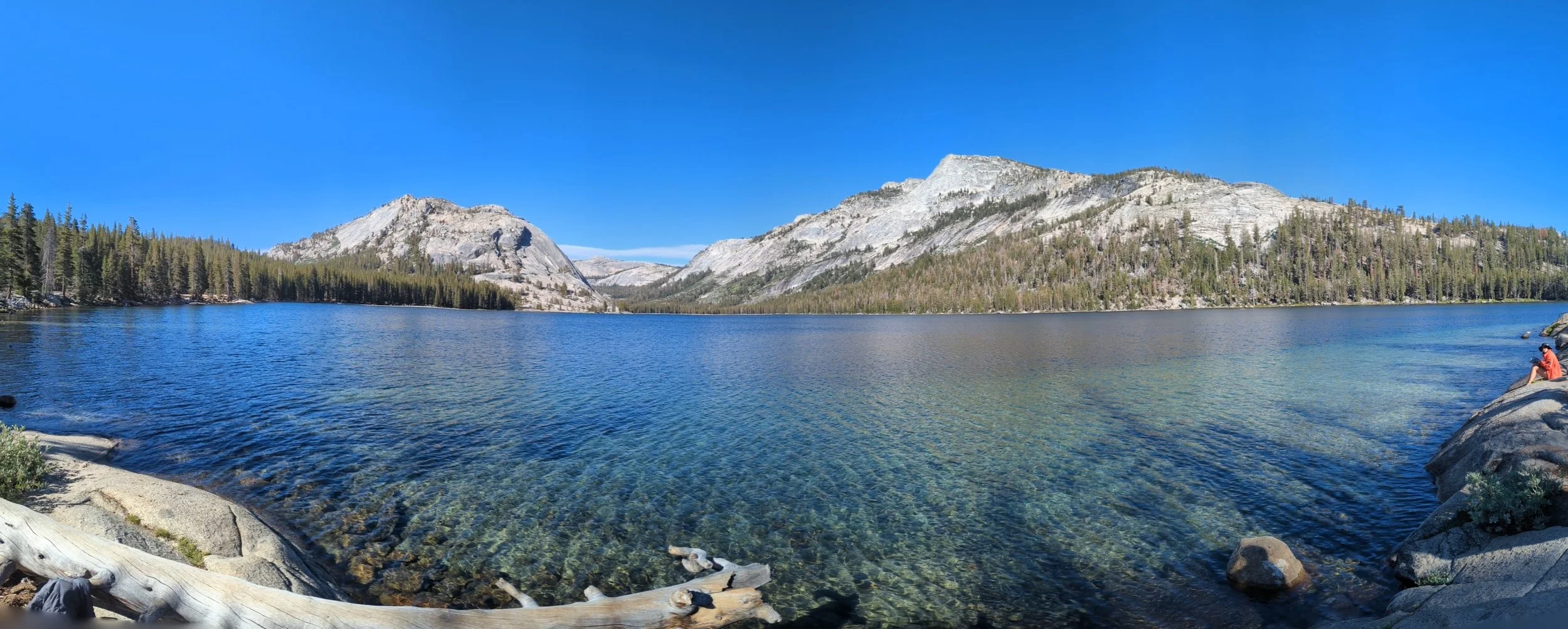 Scenic landscape of a clear blue lake surrounded by trees and mountains with a person sitting on a rock by the water.