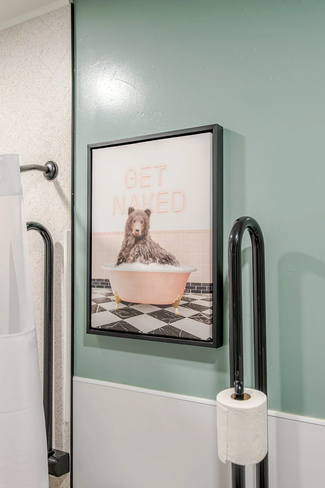 A framed sign on a green wall featuring a bear in a bathtub with the neon sign 'GET NAKED' above it.