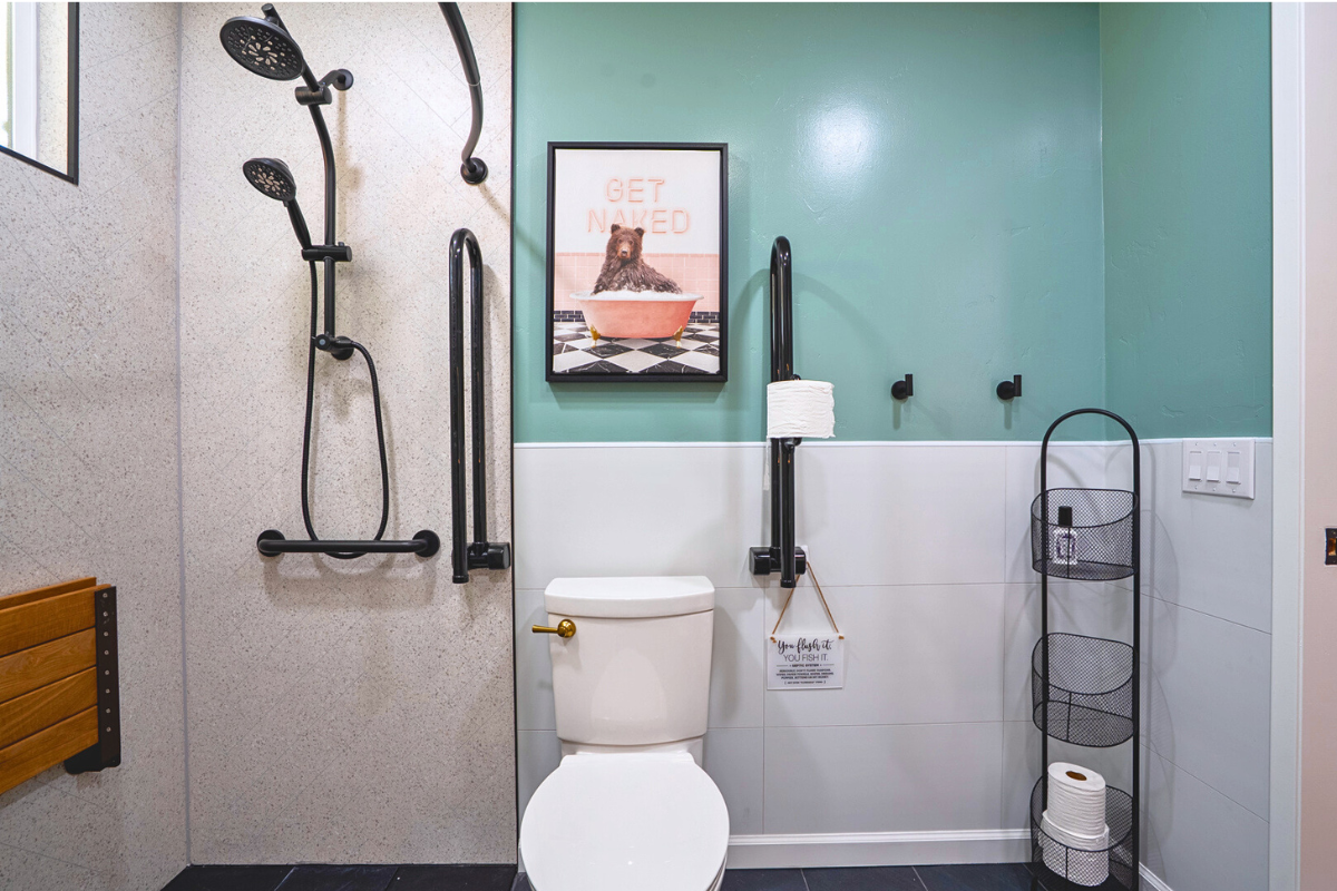 Bathroom with a toilet, grab bars, a shower area with a dual shower head, a framed bear-themed art print, a black folding grab bar with toilet paper, and a green wall with a white lower half.