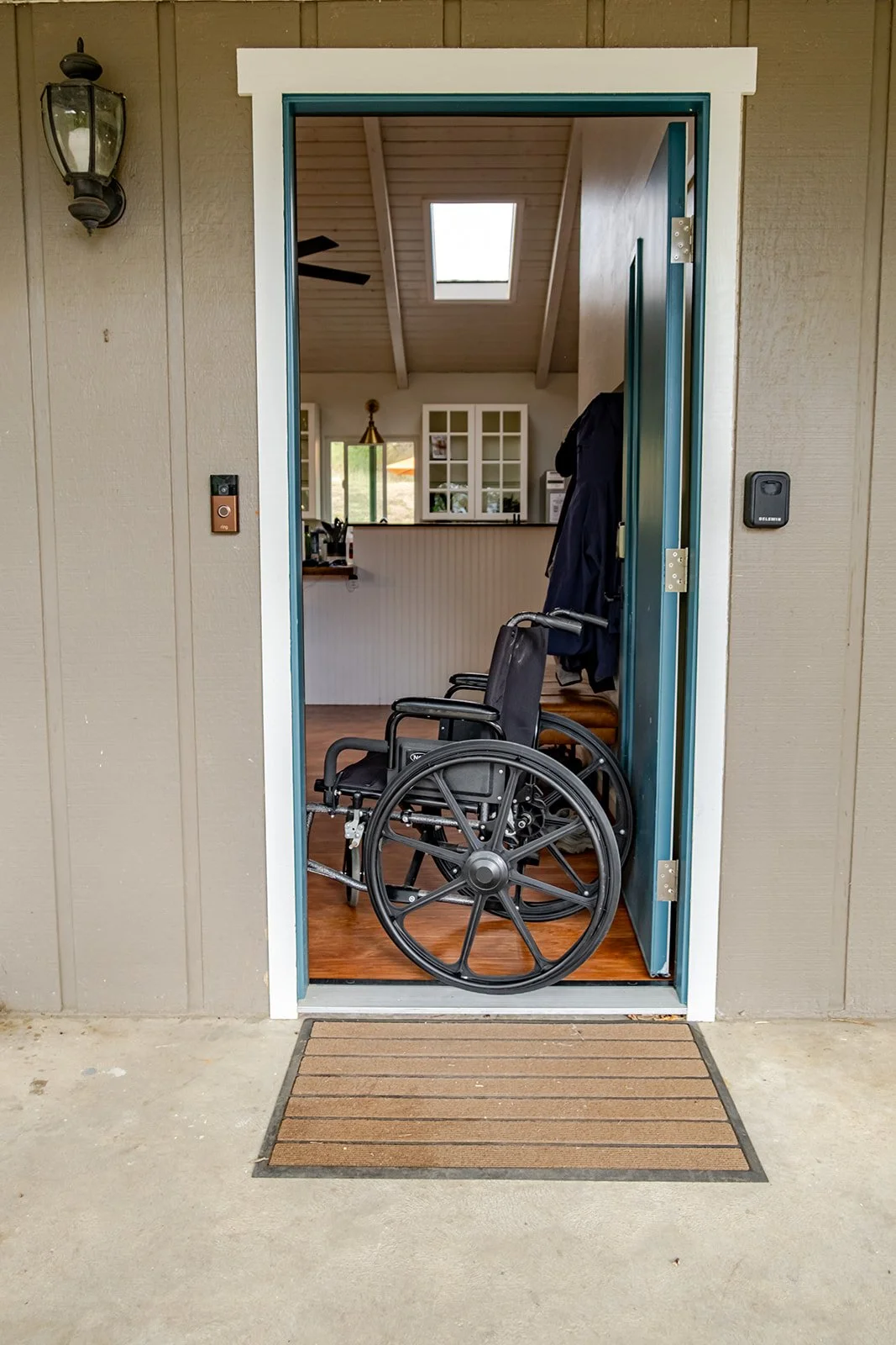 Wheelchair inside a house doorway, viewed from outside on a doormat, with a living area and kitchen in the background.