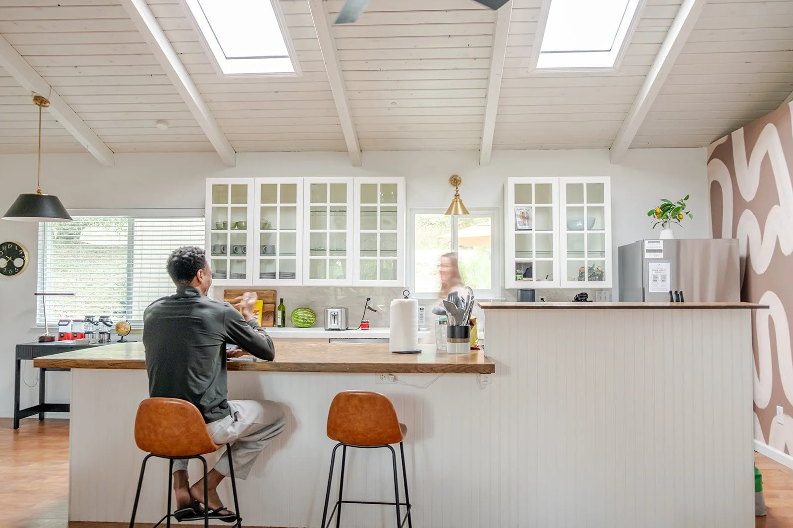A man sitting at a kitchen counter, with a woman blurred in the background, in a bright, modern kitchen with white cabinets and skylights.
