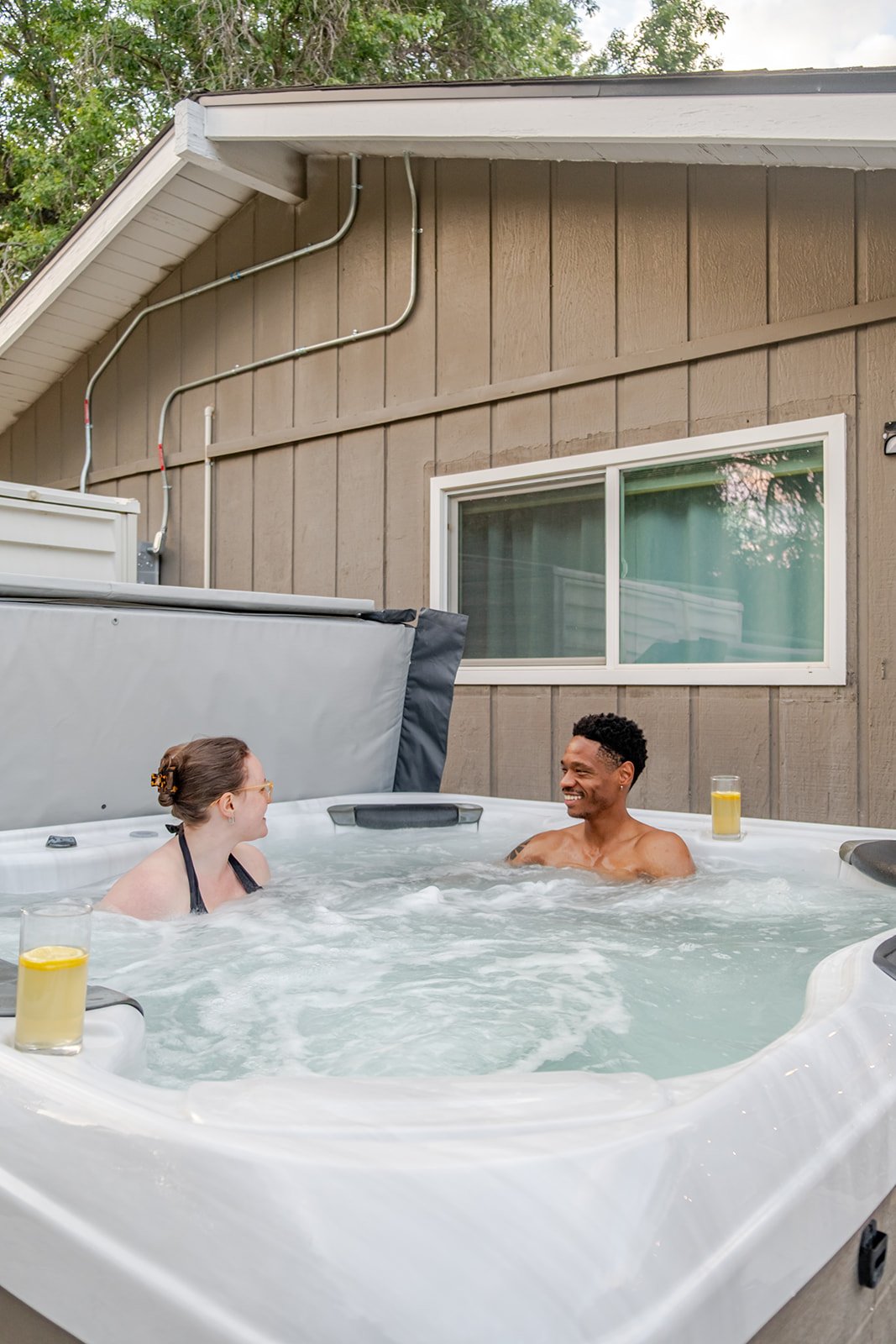Two people relaxing in an outdoor hot tub, smiling and talking, with glasses of lemonade on the edge, adjacent to a house with a window and visible exterior pipes.
