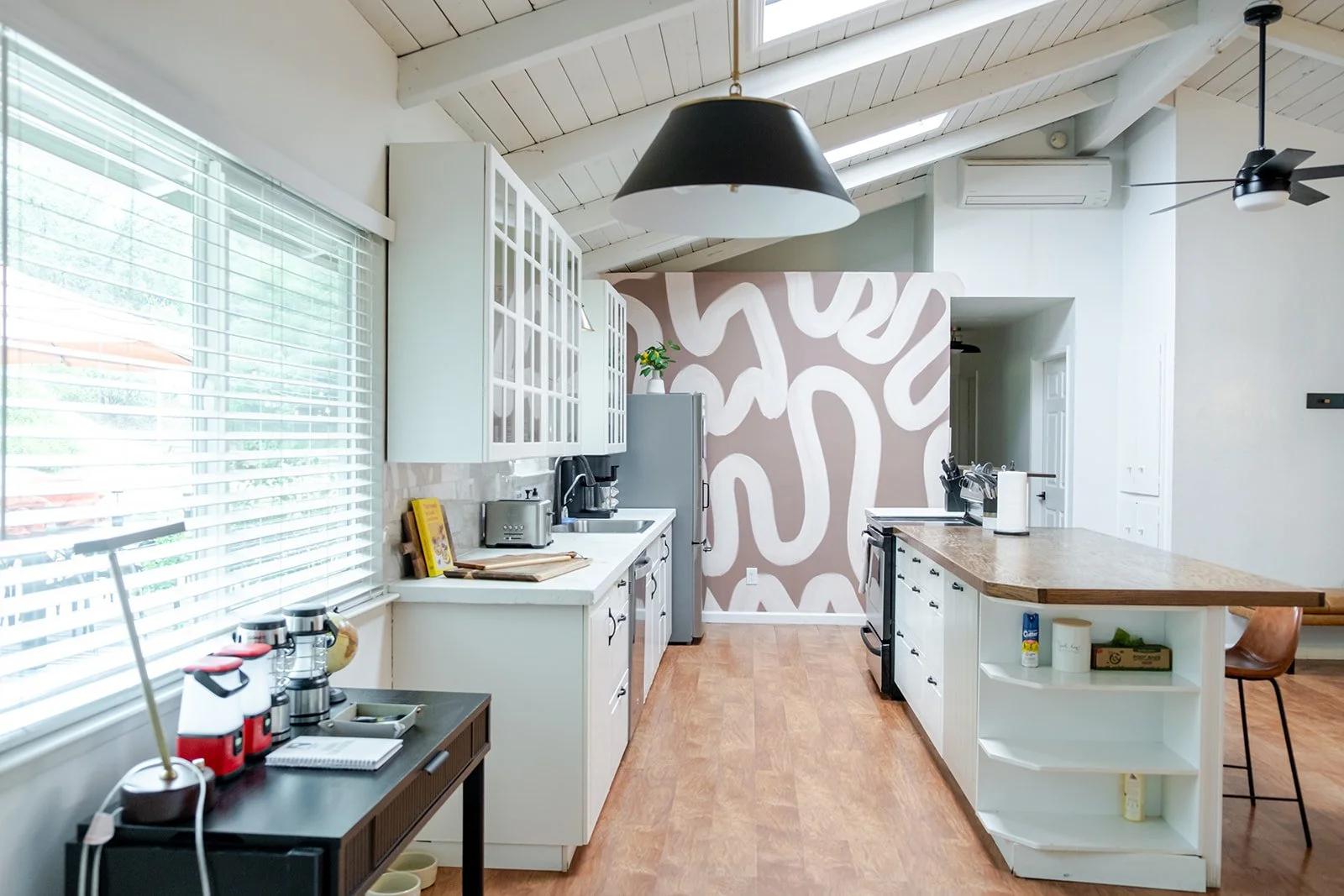 Modern kitchen with white cabinets, a wooden island, patterned pink and white accent wall, large window with blinds, and various small appliances.