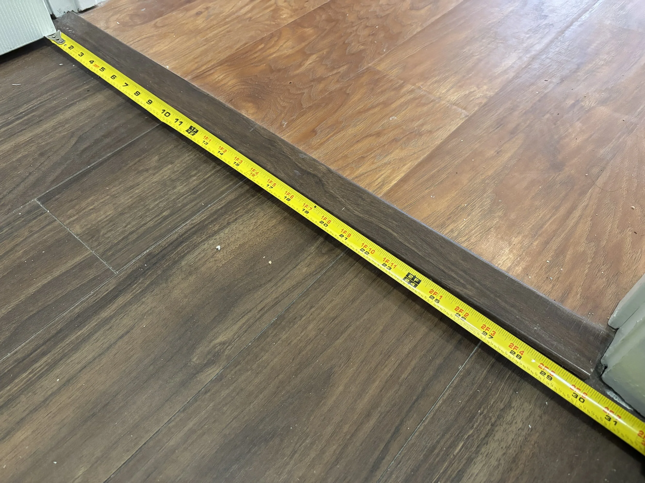 Close-up of the edge of a wooden floor with a yellow measuring tape extending along it, showing measurements in inches.