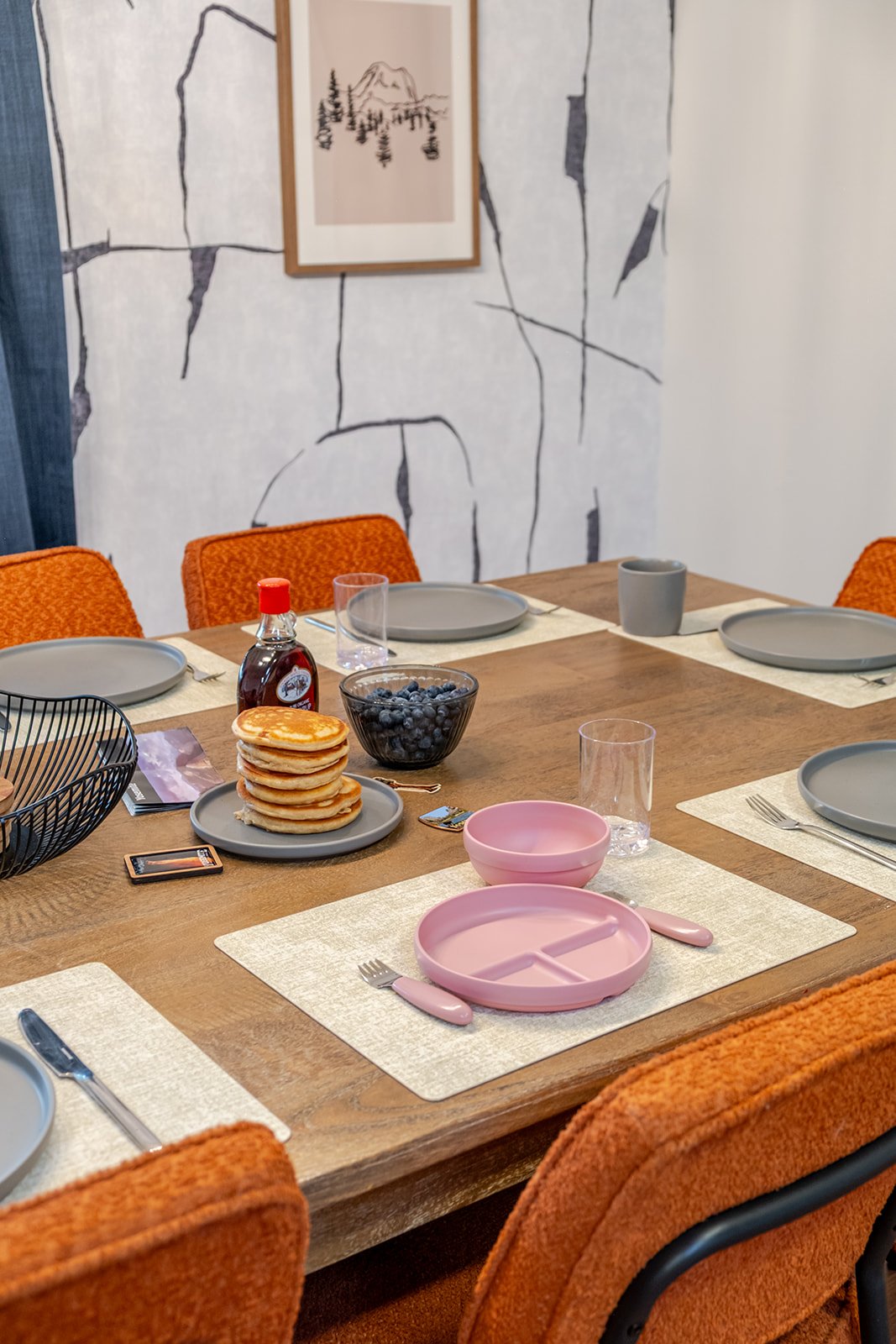 A dining table set with plates, glasses, a bowl of blueberries, a stack of pancakes, and a pink divided plate with a spoon and fork. Orange upholstered chairs surround the table, with a wall hanging of a mountain and trees in the background.