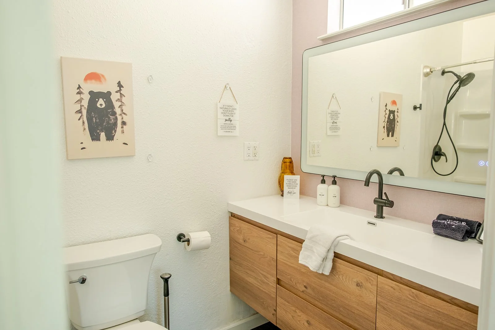 A bathroom with a white toilet, a wooden vanity with a white countertop, a large mirror, and a shower with a hand-held showerhead. Decor includes a bear and sun art on the wall, signage, and toiletries.