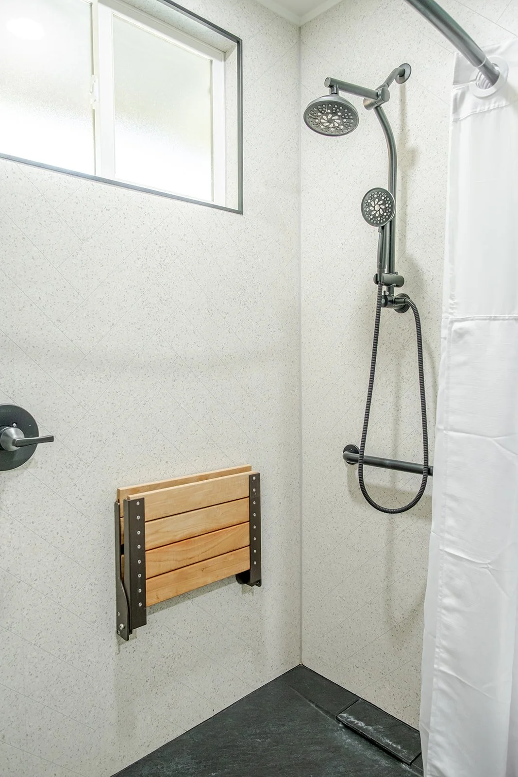 A shower with a showerhead, a window, a wooden foldable shower seat, and a white shower curtain in a minimalist bathroom.