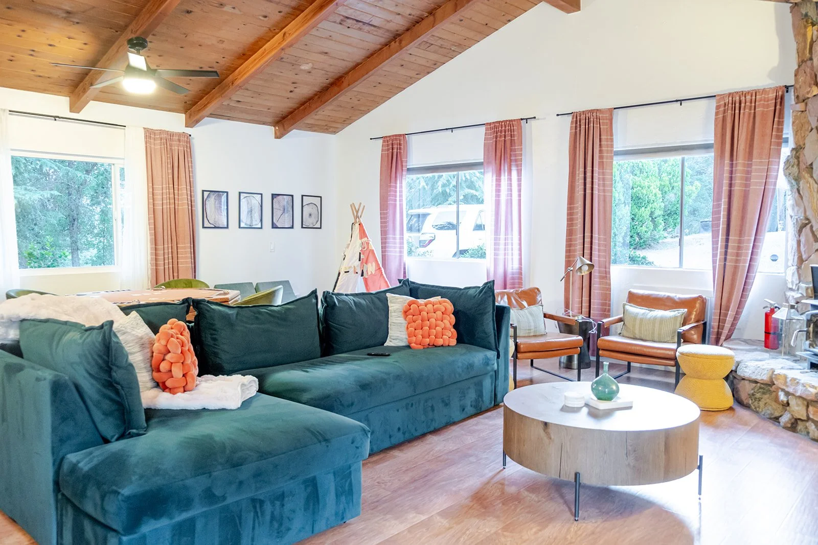 Living room with teal sectional sofa, orange cushions, wooden ceiling with beams, windows with pink curtains, leather armchairs, a round coffee table with a green vase, artwork on the wall, and a stone fireplace.