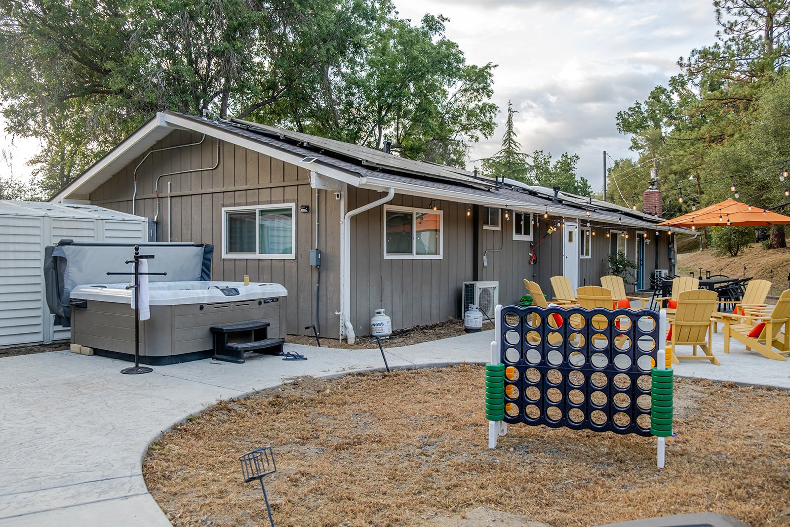Backyard patio with a brown house, a hot tub, a Connect Four game, Adirondack chairs, a patio umbrella, string lights, and trees in the background.