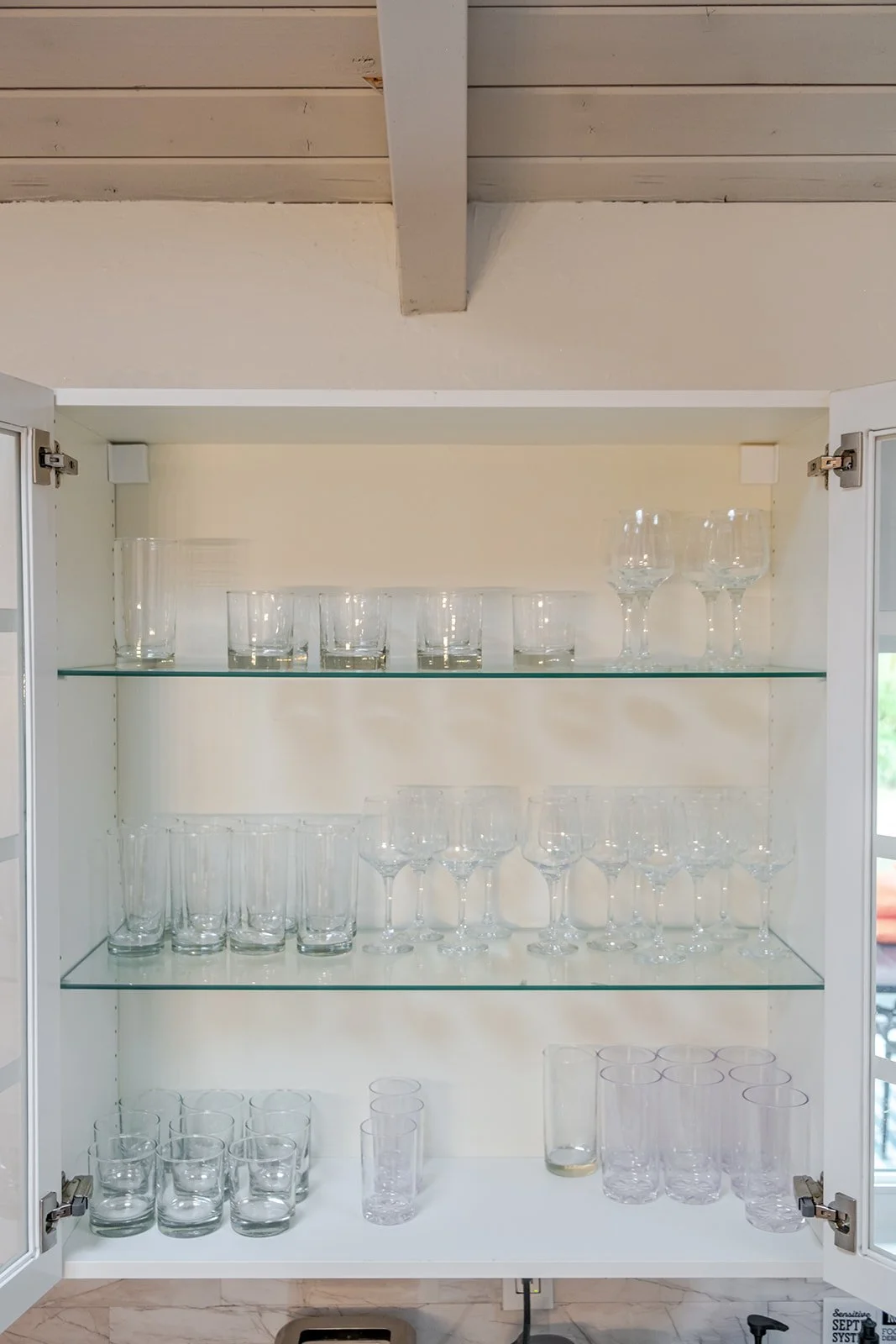 White glass cabinet with three shelves filled with various clear glassware, including glasses and wine glasses.