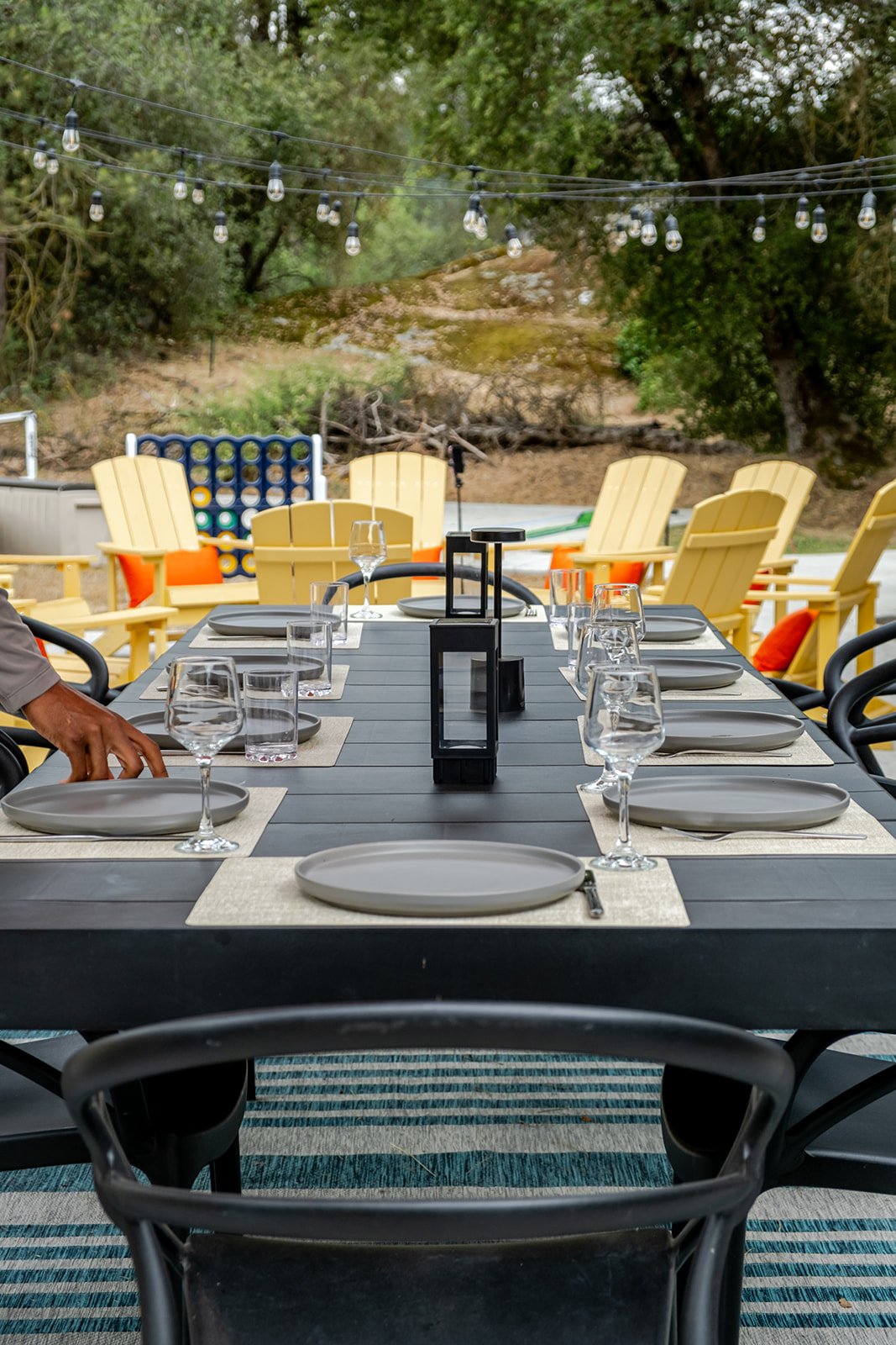 Outdoor dining table set for a meal with plates, glasses, and a lantern centerpiece, surrounded by yellow Adirondack chairs and string lights, with trees in the background.