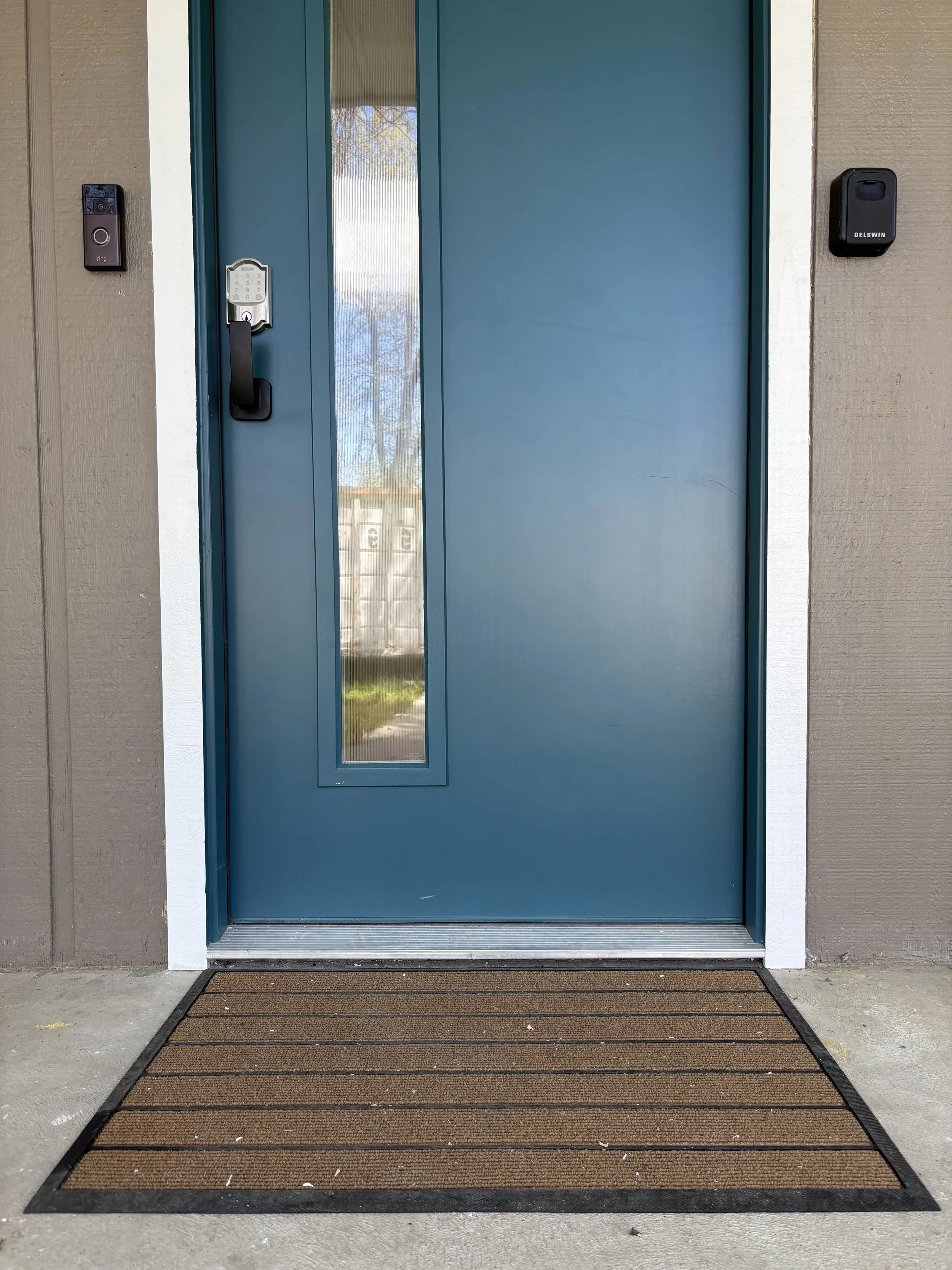 Blue front door with a narrow window on the left, with an electronic lock and a keypad. There is a welcome mat in front of the door on a concrete porch.