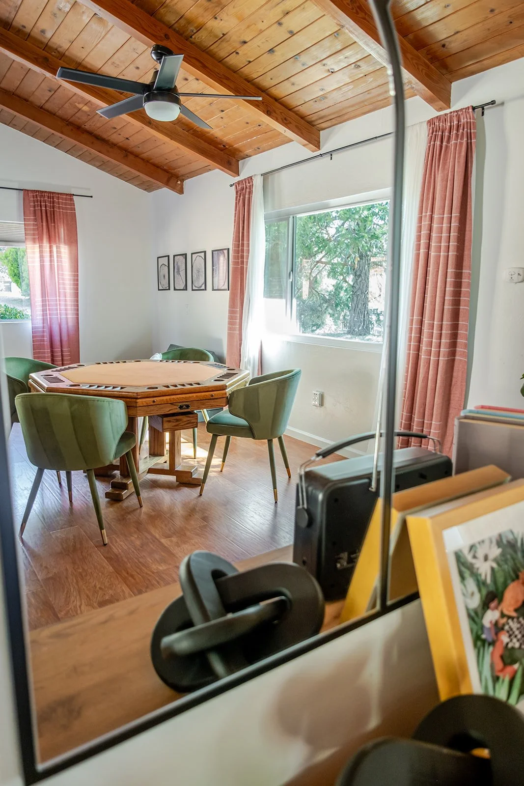A dining room with a wooden ceiling and hardwood floor, featuring a round green table surrounded by five white and green chairs, large windows with pink and white curtains, and framed artwork on the wall.