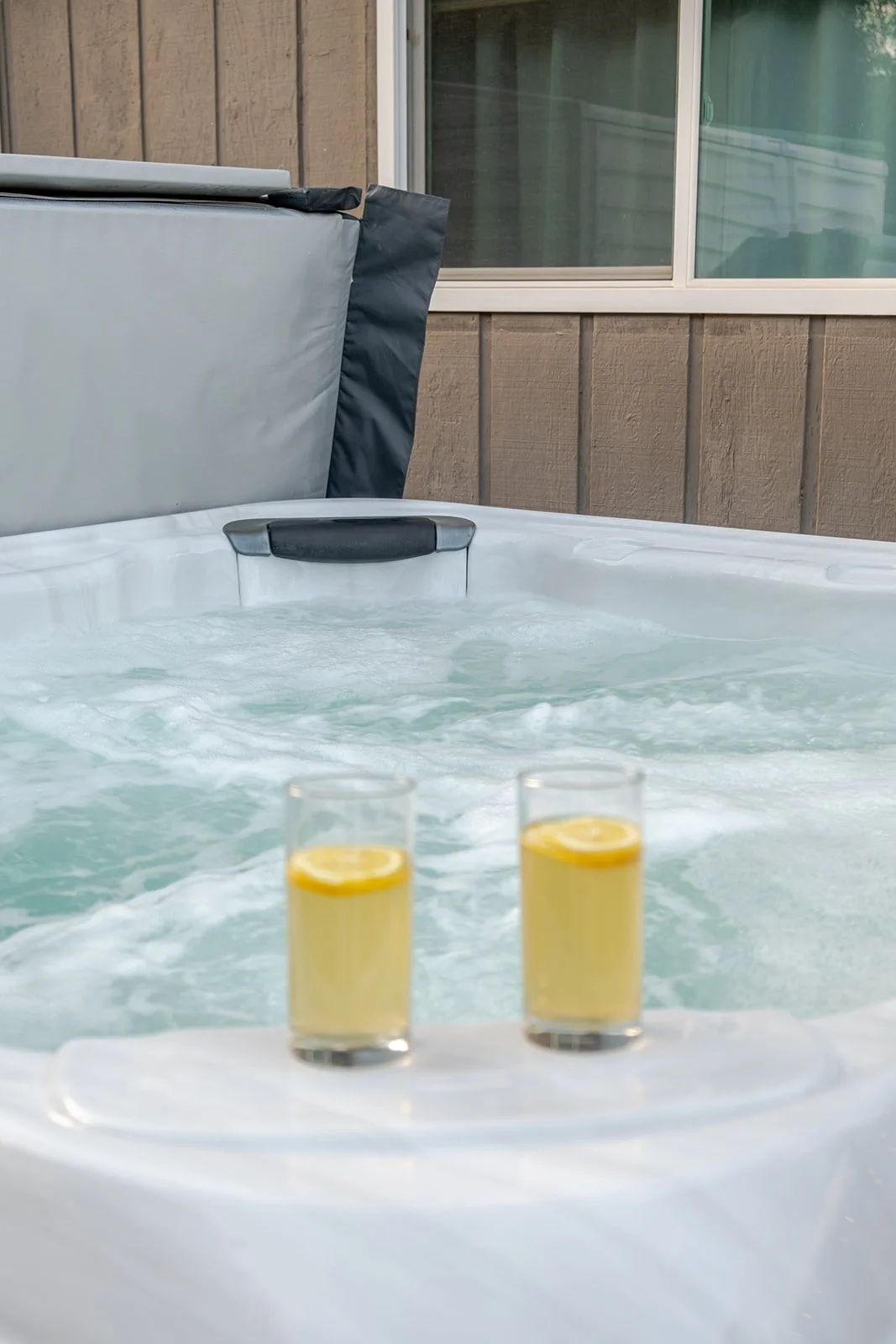 Two glasses of lemonade with lemon slices on a tray near a hot tub with water jets, on a balcony with a window and wooden paneling.