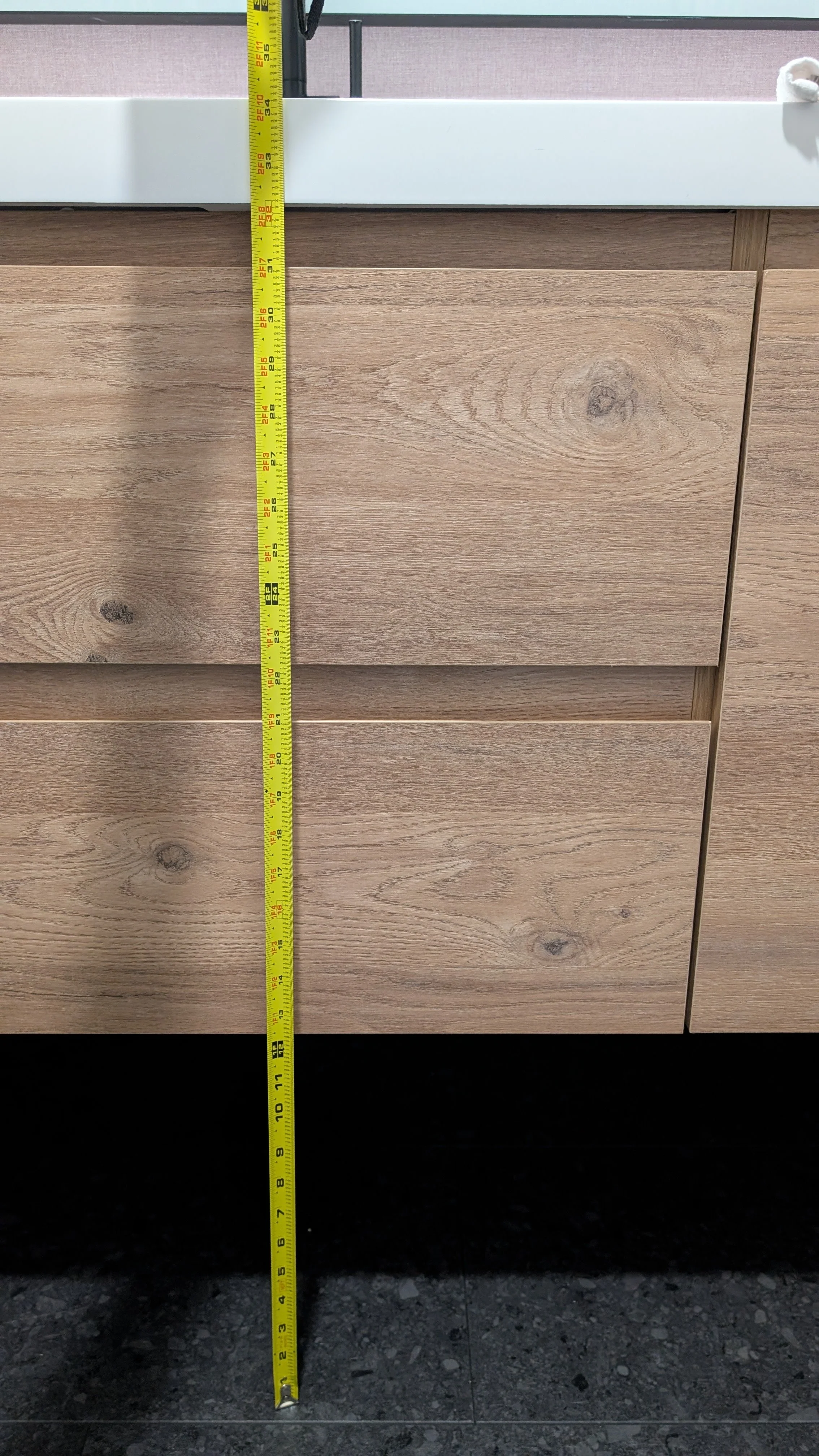 A measuring tape standing vertically in front of light-colored wooden drawers, with a white countertop above, on a dark speckled floor.