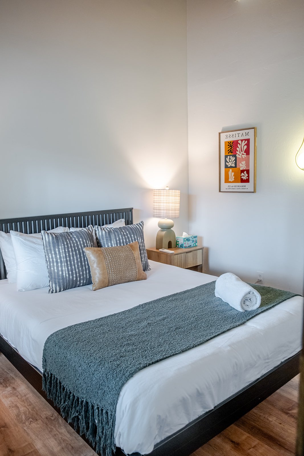 A neatly made bed with white sheets, gray and beige decorative pillows, and a dark gray bed runner in a cozy bedroom. A rolled white towel is placed on the bed. Next to the bed is a wooden nightstand with a modern white lamp, a tissue box, and a remo