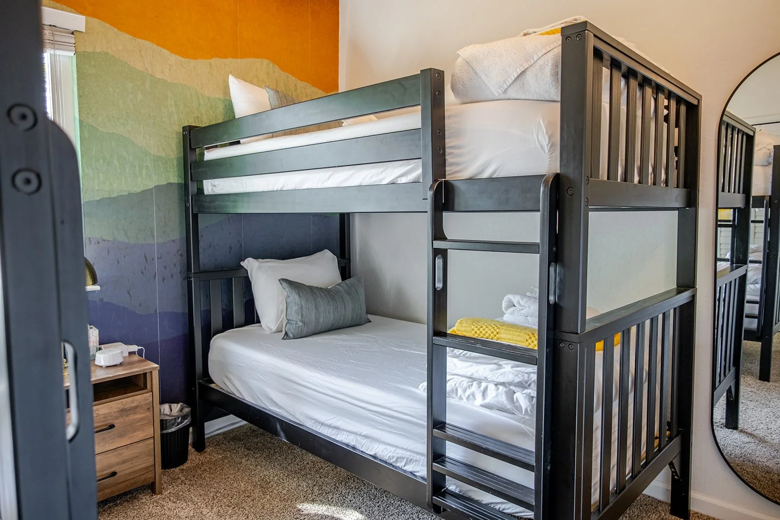 Bunk beds with black metal frame, beds made with white sheets and pillows, small wooden nightstand with a lamp and a disposable cup, colorful wall with painted landscape design, rug on beige carpeted floor, reflection in mirror on the right side.