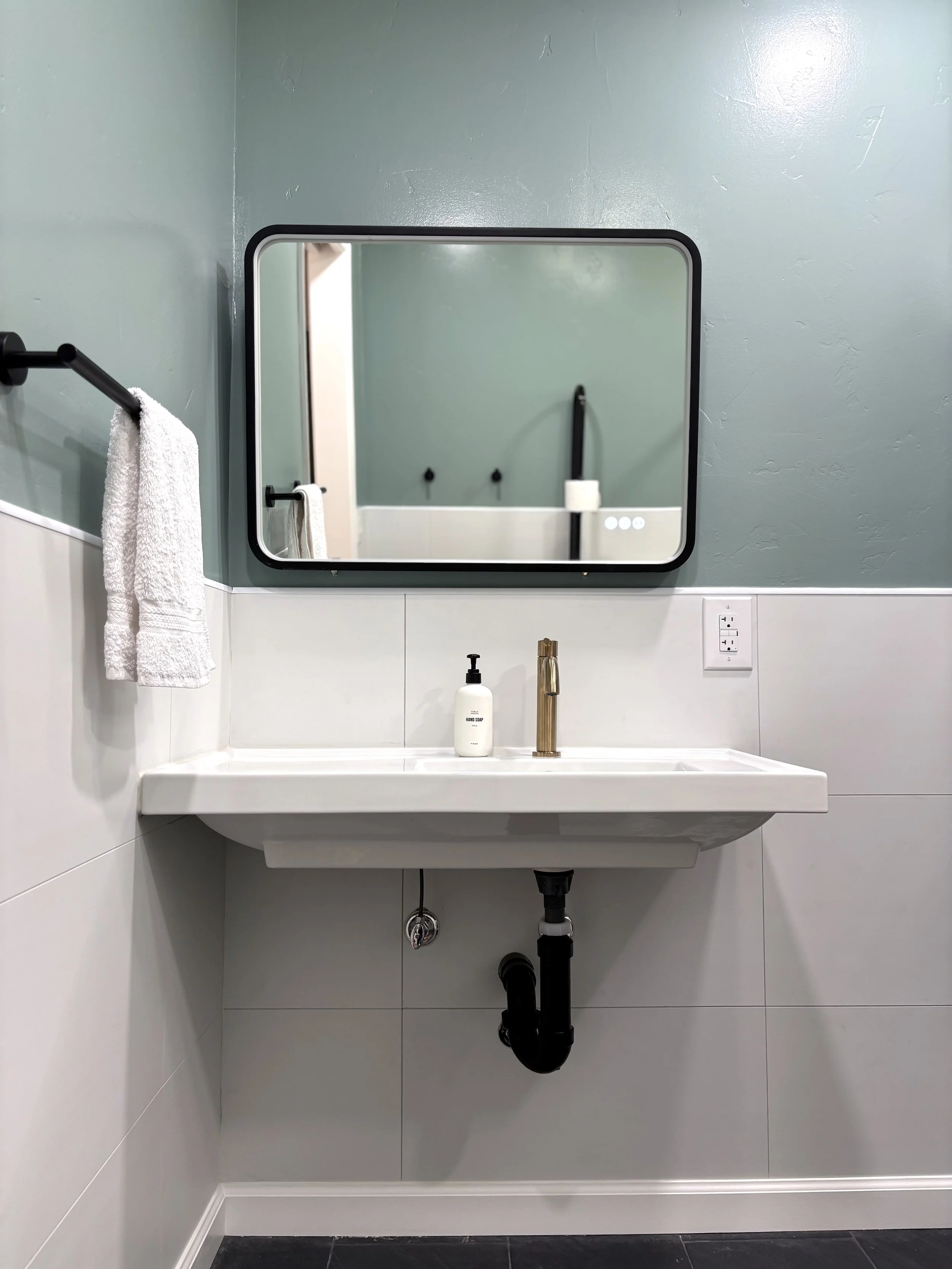 A bathroom sink with a white rectangular basin, a gold faucet, a soap dispenser, a mirror, a towel on a black towel bar, and a wall outlet, with visible plumbing underneath.