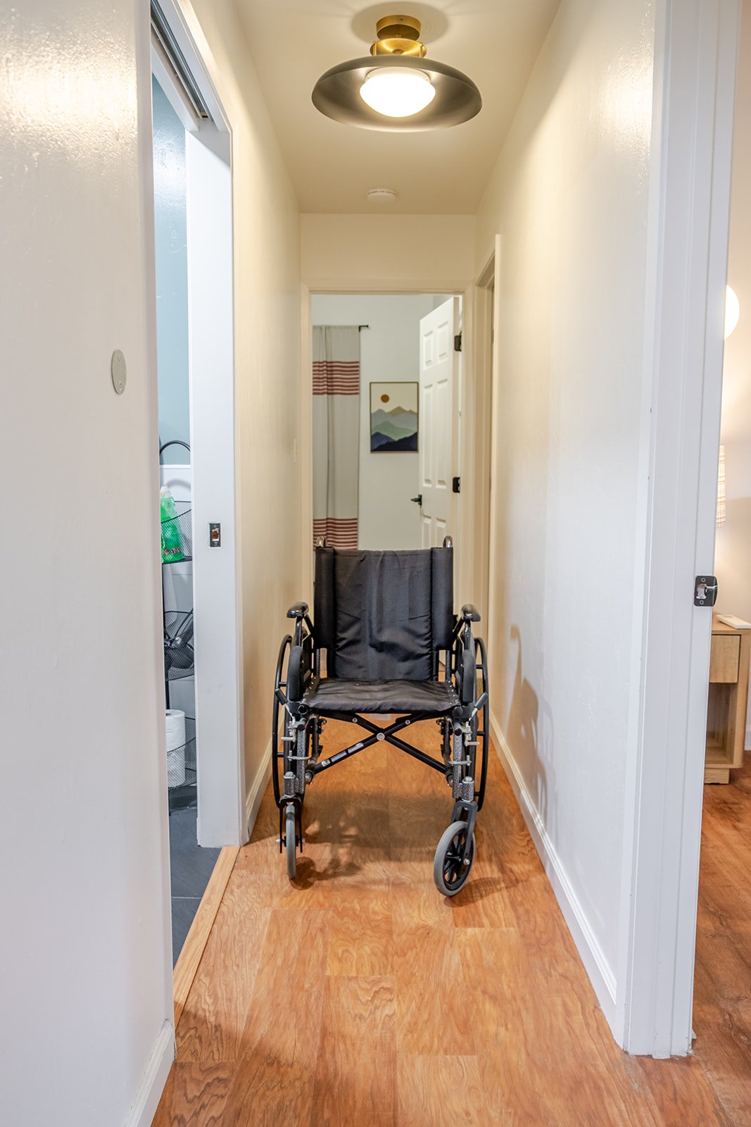 Hallway with a black wheelchair positioned in the center, white walls, wooden flooring, ceiling light fixture, and an open door at the end leading to another room with artwork and curtains