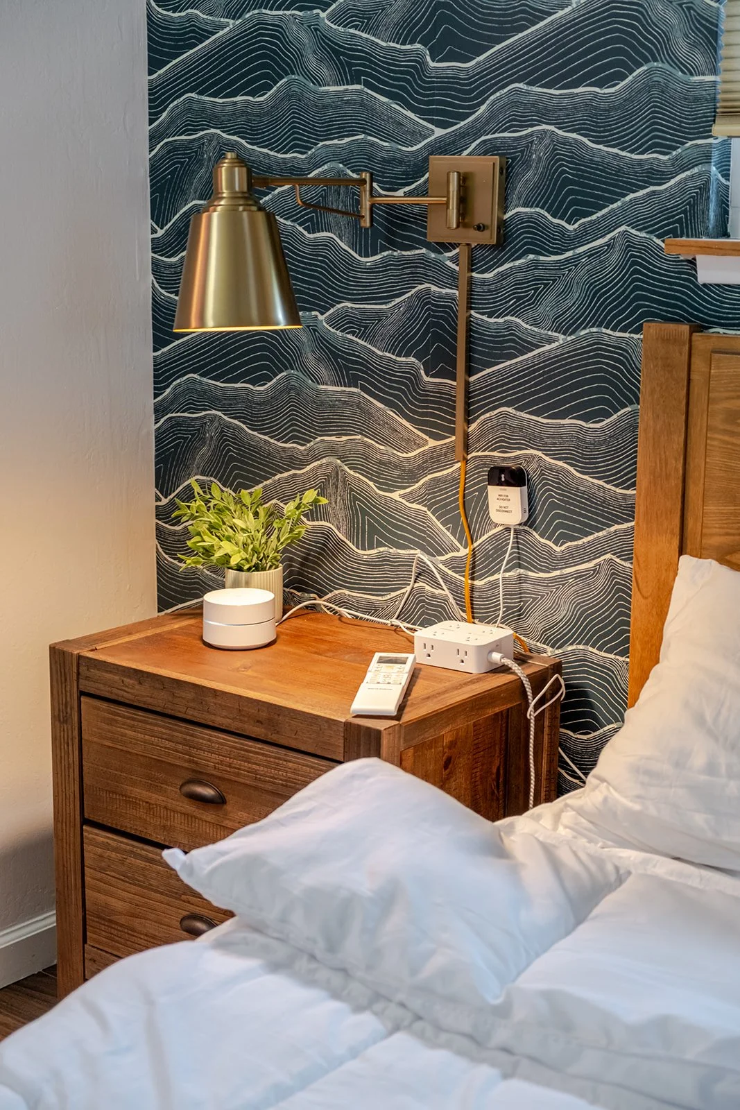 A bedroom nightstand with a potted plant, a white smart device, a remote, and a power strip. A brass wall-mounted lamp is above the nightstand, and a blue mountain-patterned wallpaper decorates the wall. Part of a bed with white bedding and a pillow 
