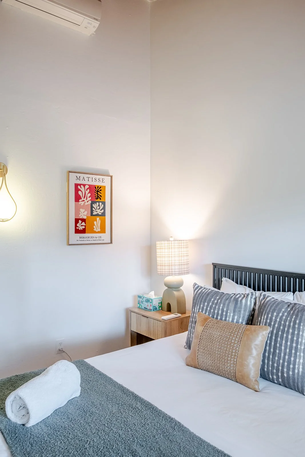 A neatly made bed with striped and beige pillows, a rolled towel at the foot, a wooden nightstand with a lamp, tissue box, and remote control, a framed Matisse artwork on the wall, and part of an air conditioning unit in a bright, modern bedroom.