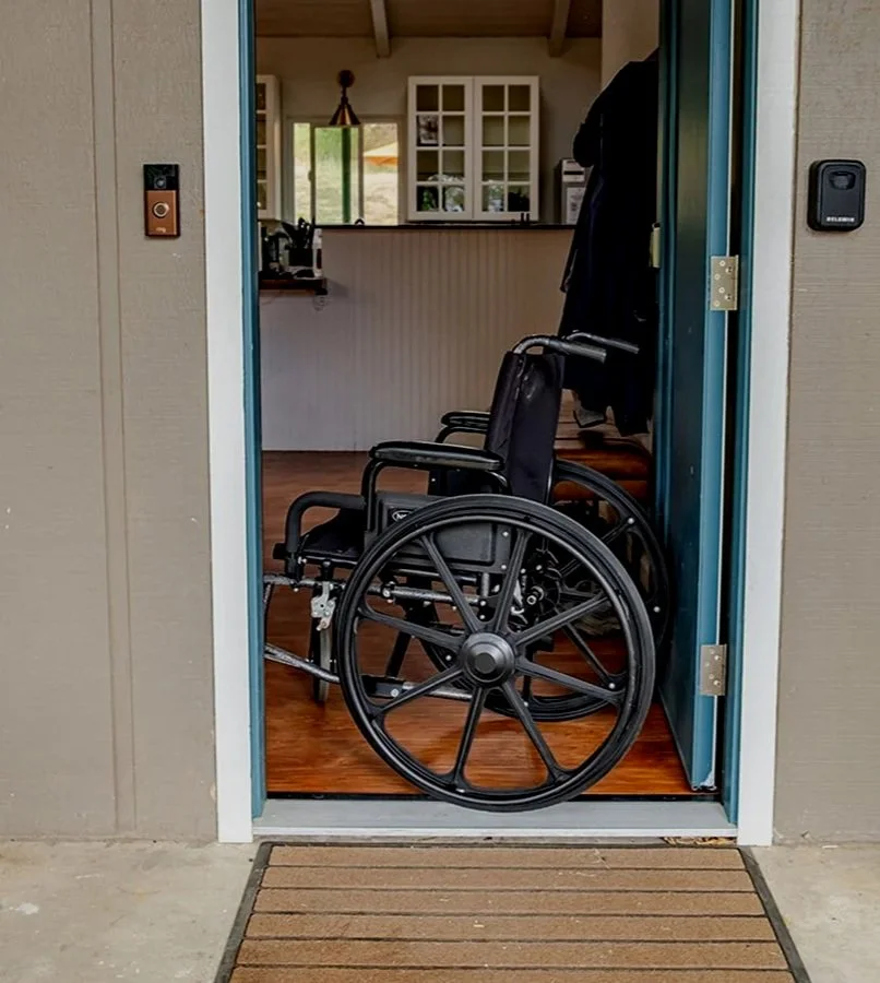 A black wheelchair sits in the threshold of the front door to demonstrate zero-step entrance into the house. Inside the house you can see the kitchen.
