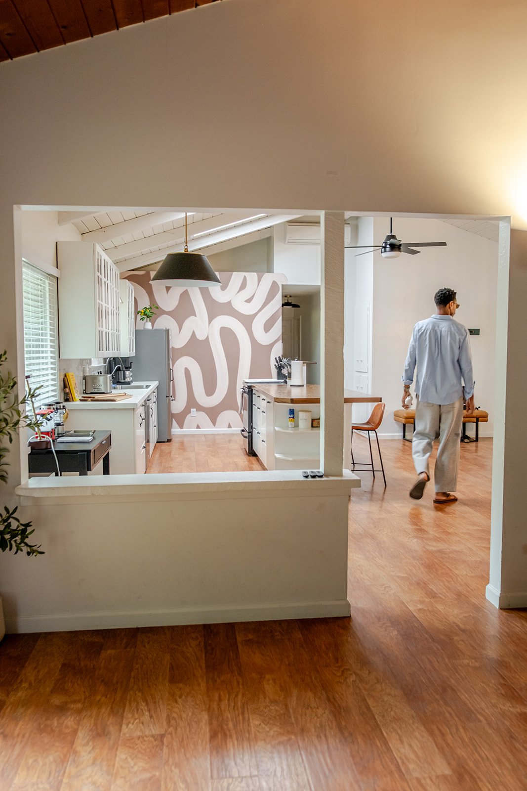 A man walking in a modern kitchen with white cabinetry, a black pendant light, and a wall with an abstract brown and white pattern. The kitchen has wooden floors and a ceiling fan.