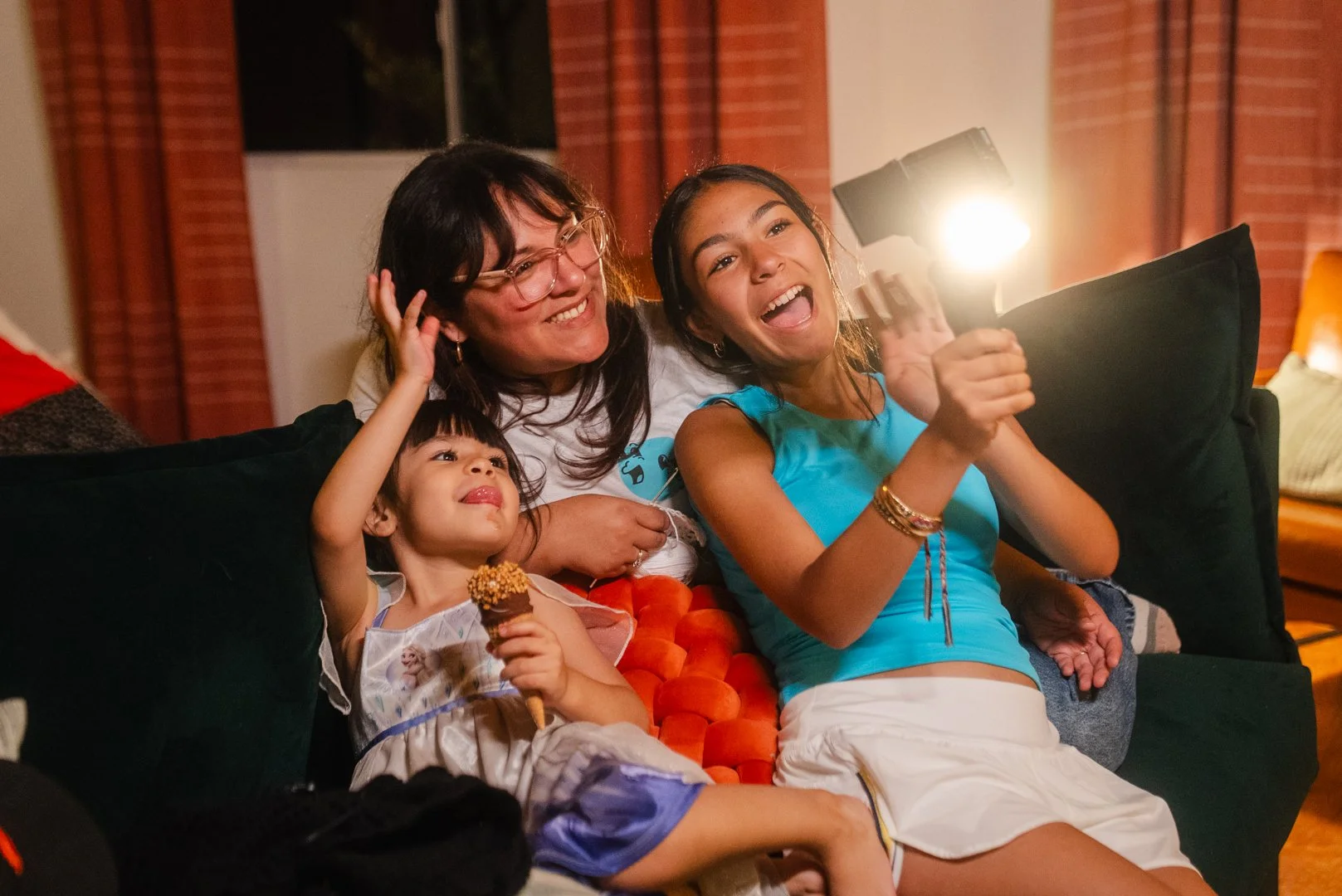 A group of four people, including two young girls and two women, sitting on a couch, smiling, and taking a selfie with a flashlight, in a warmly lit living room with brick walls and a window.