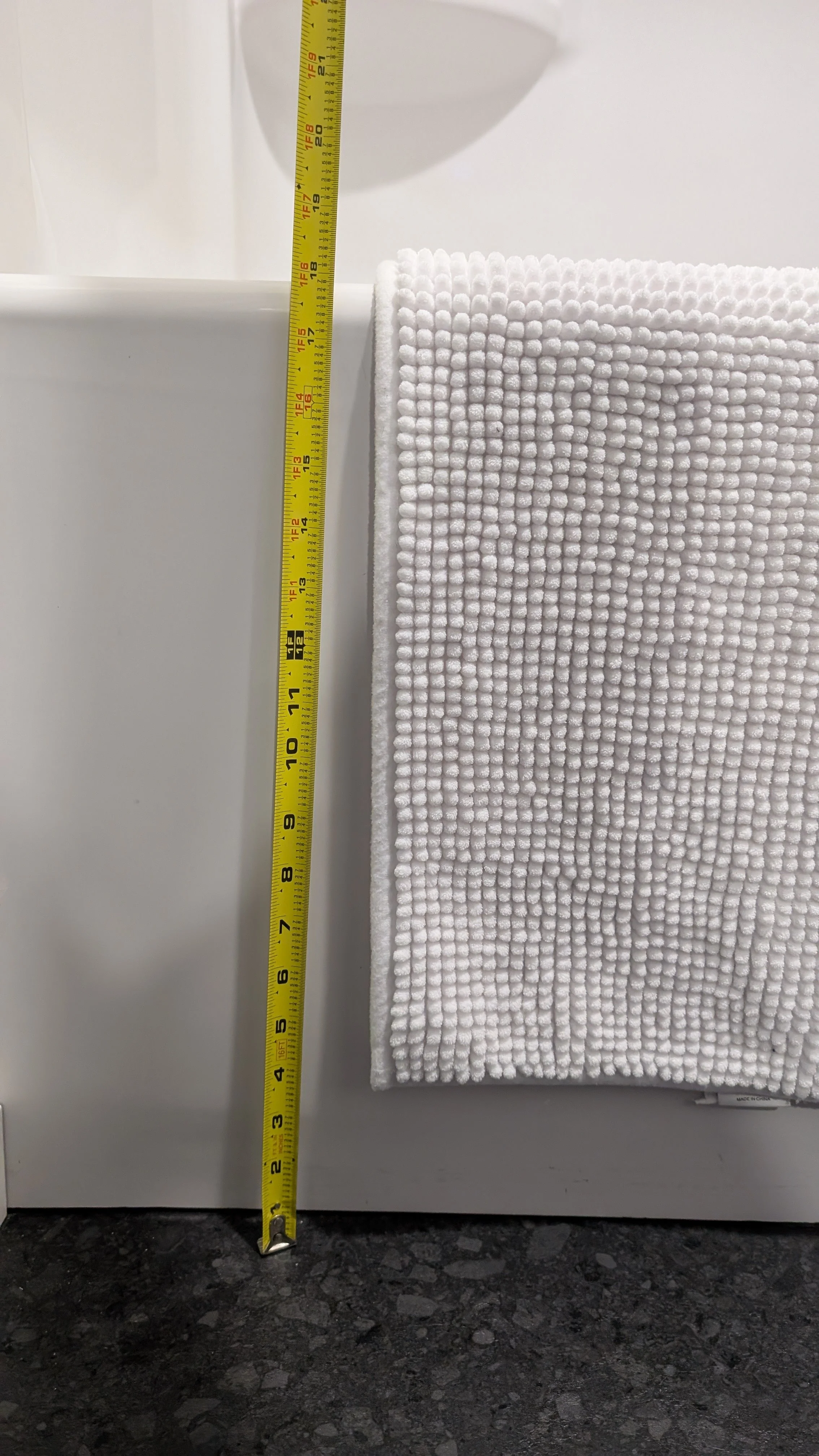A yellow measuring tape standing vertically next to a white towel hanging with a textured pattern. The measuring tape shows a height of approximately 16 inches. The background consists of a white wall and a bathroom countertop with a black and gray s