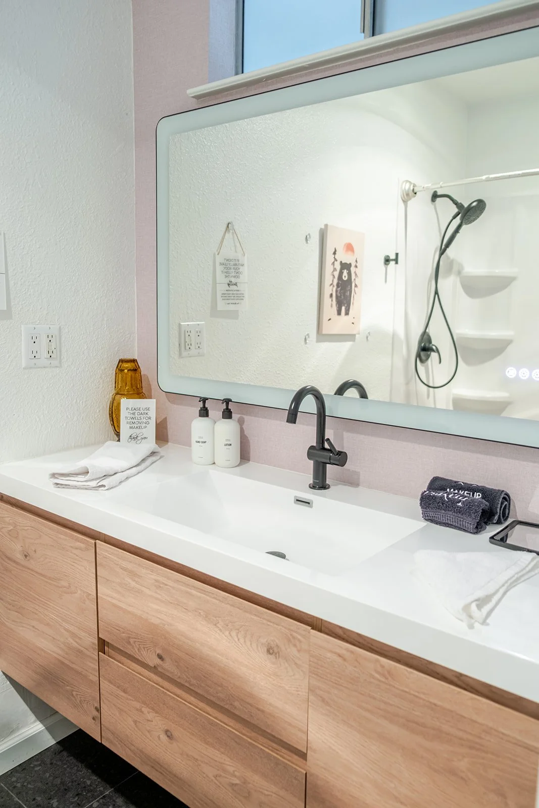 A modern bathroom sink area with a large mirror, light wood vanity, black faucet, two white soap dispensers labeled 'Hand Soap' and 'Lotion,' a rolled dark towel on a small stand, and decorative items including a amber glass vase and a small sign. Th